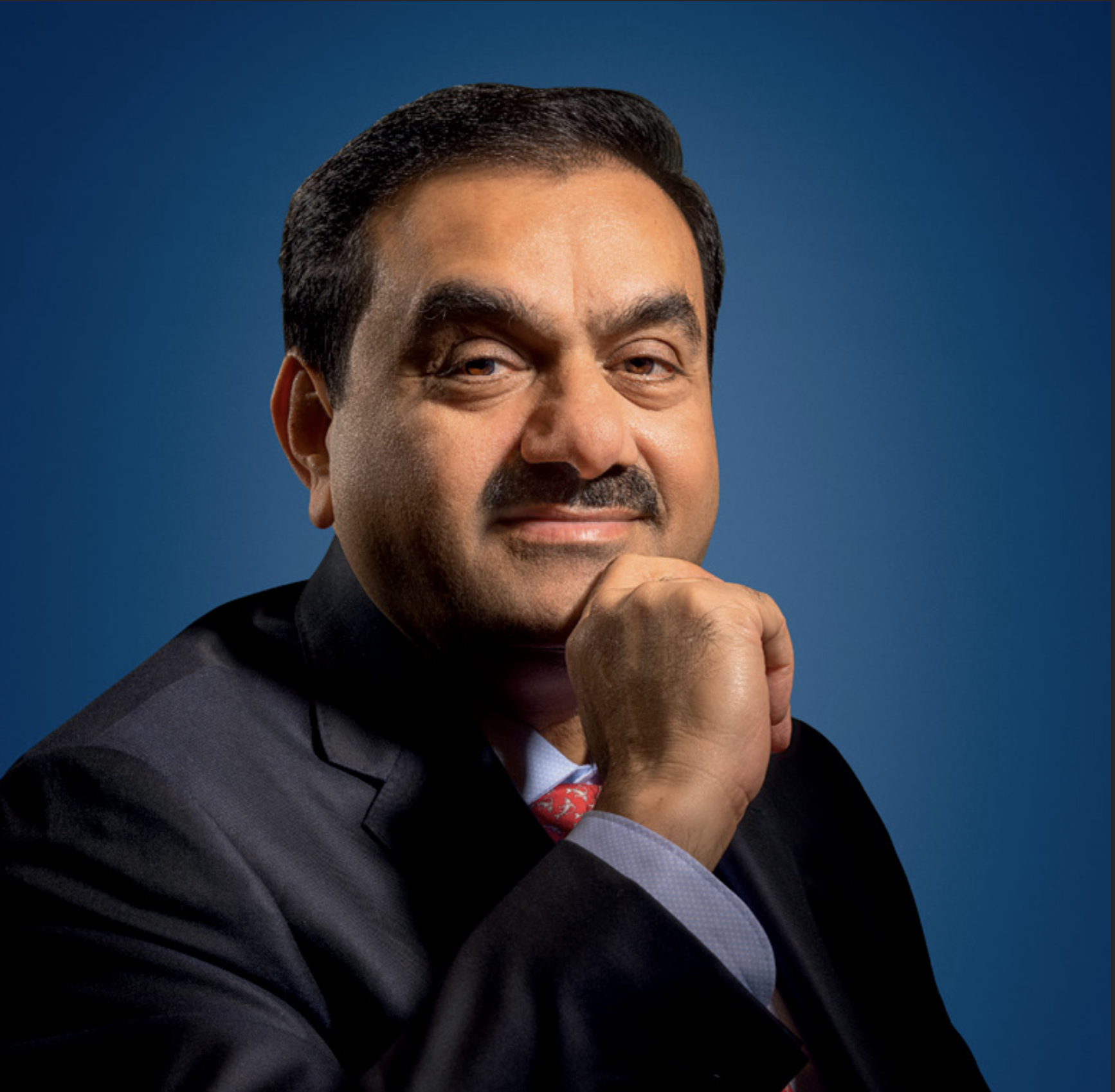A headshot of Gautam Adani, a smiling man in a suit, with his hand under his chin.