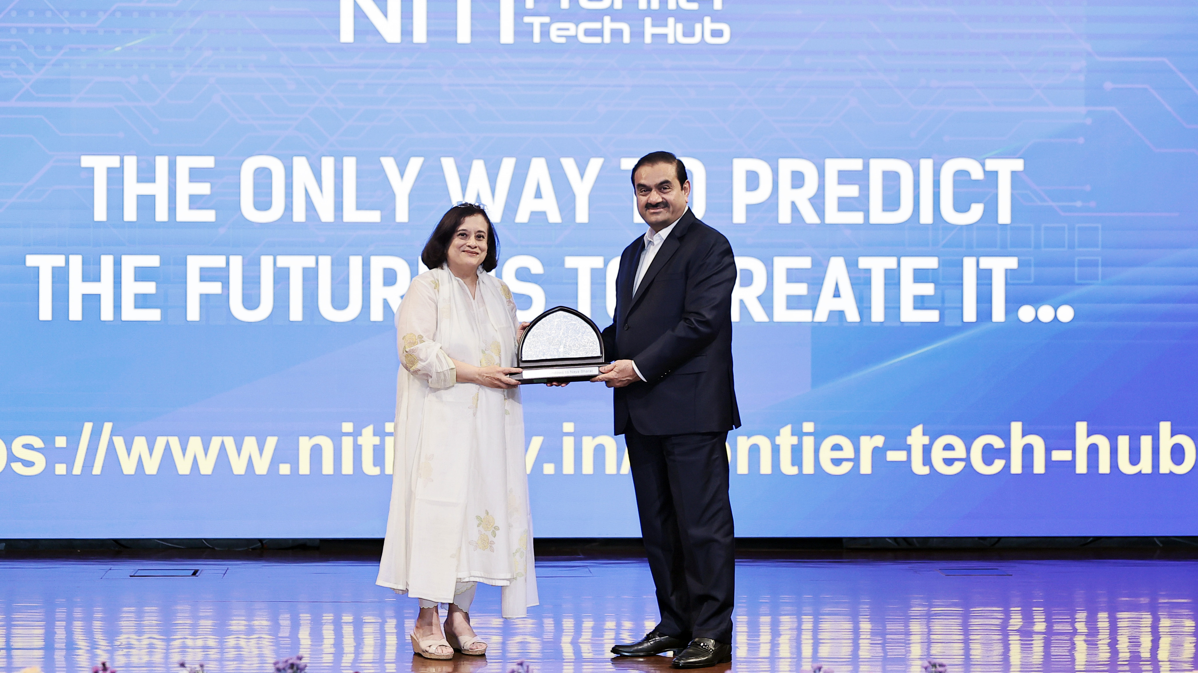 Two individuals, a man and a woman, are on stage exchanging an award in front of a large screen displaying a tech-related quote.