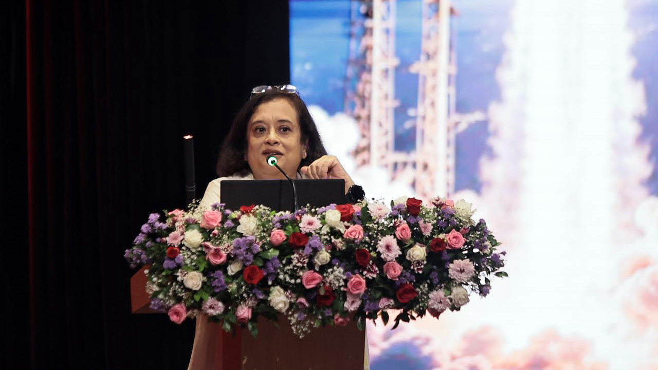 A woman speaks at a flower-adorned podium with a rocket launch displayed behind her.