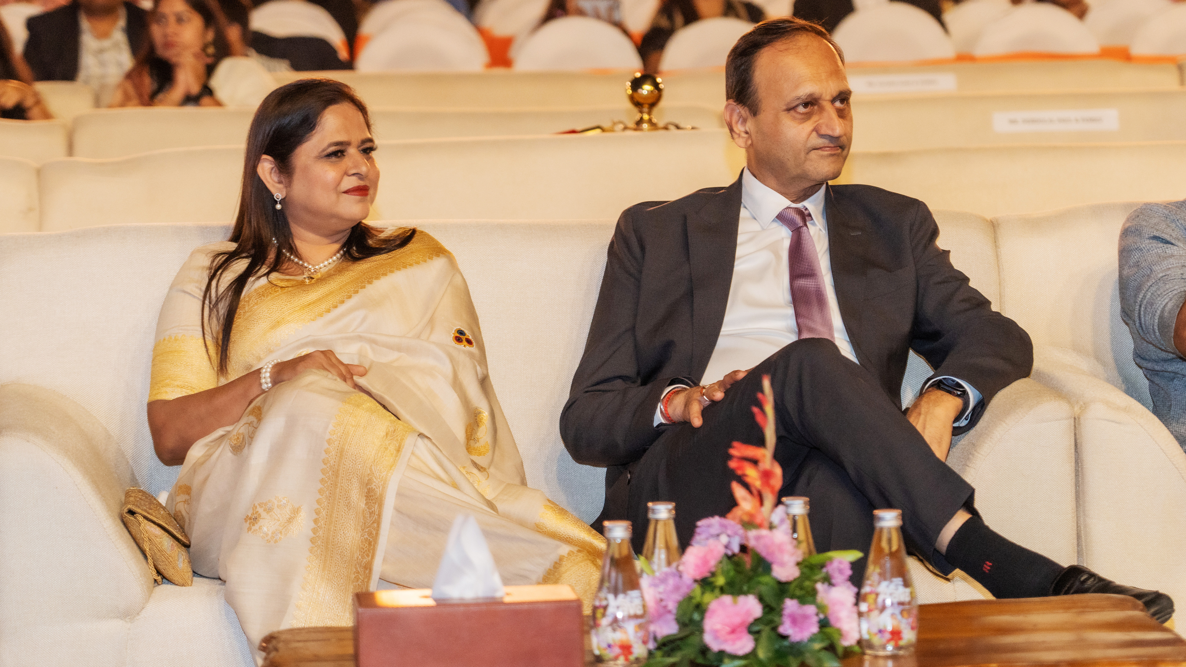 Two formally dressed individuals, a woman in a saree and a man in a suit, seated in an audience.