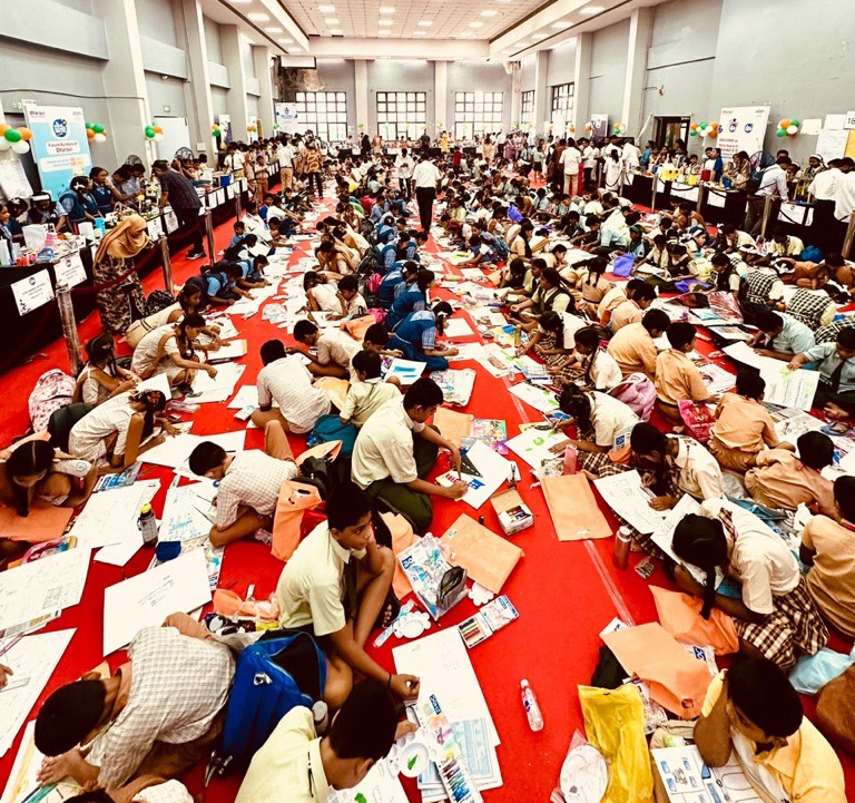 Many children sit on a red carpet in a large hall, engrossed in drawing and coloring during an event.
