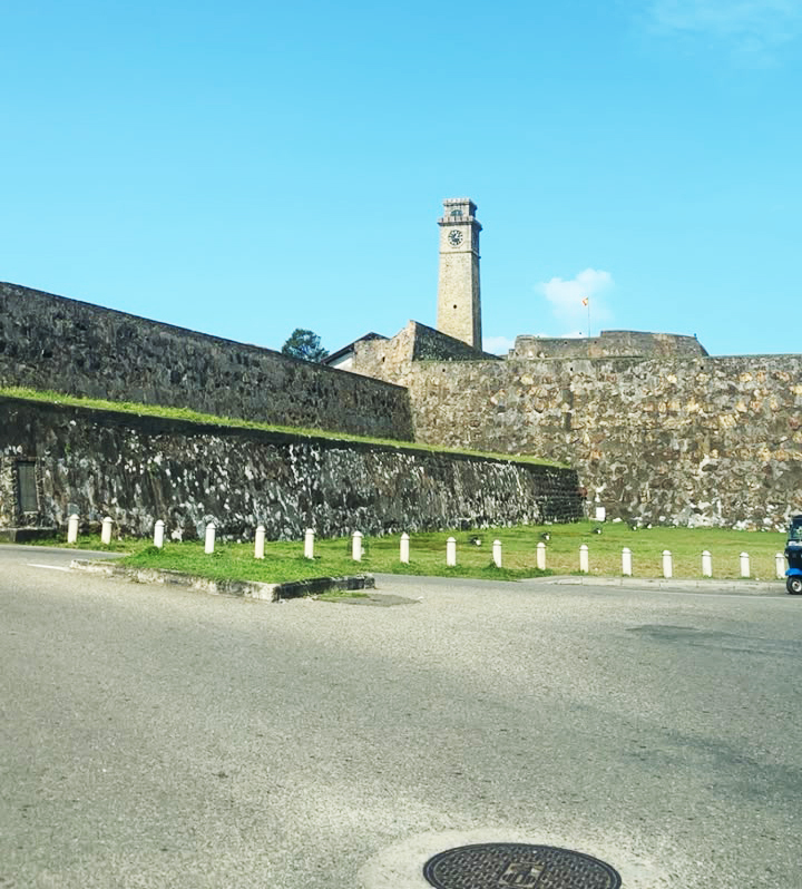 Galle Fort clock tower rises above ancient stone walls against a clear blue sky, with a road in the foreground.