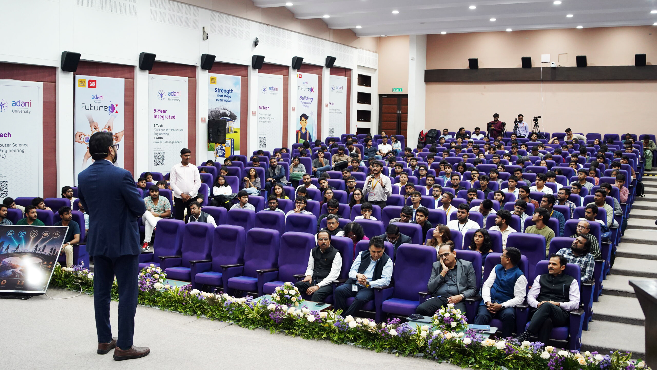 A speaker addresses a large audience seated in purple chairs in a university auditorium.