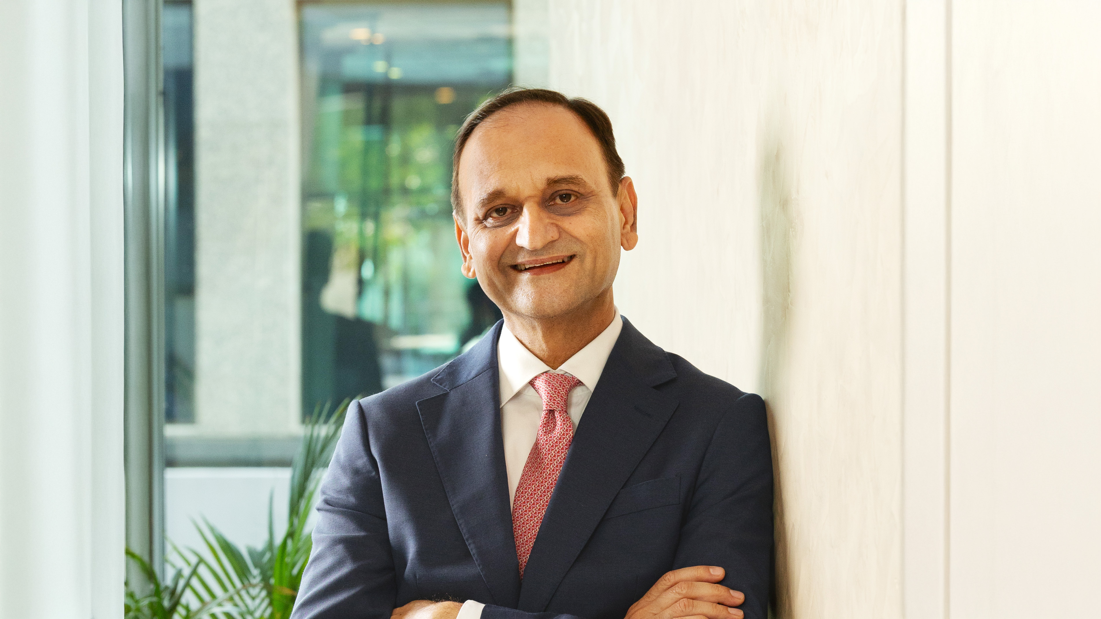 Headshot of a smiling businessman in a navy suit and red tie, leaning against a wall.