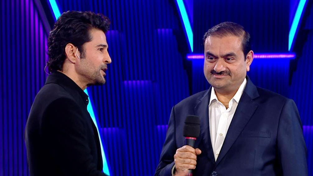 Two men, one in a black jacket and one in a blue suit holding a microphone, converse on a stage.