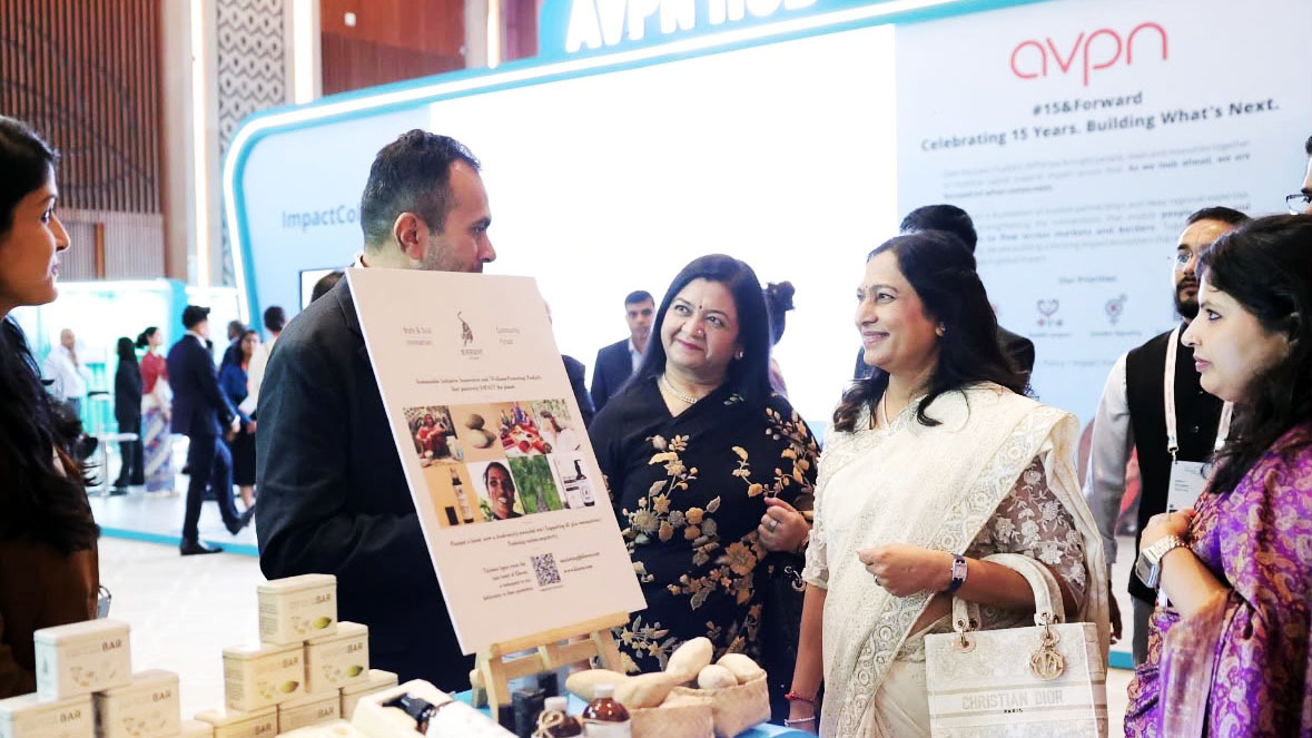 Man presents a product display board to three women at an event booth with "AVPN HUB" branding in the background.
