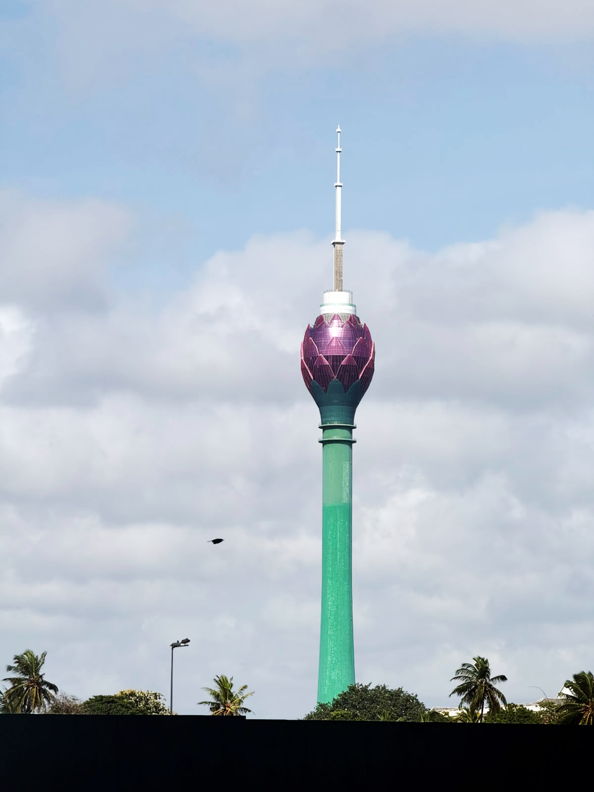 A tall green and pink Lotus Tower stands against a cloudy sky, with palm trees in the foreground.