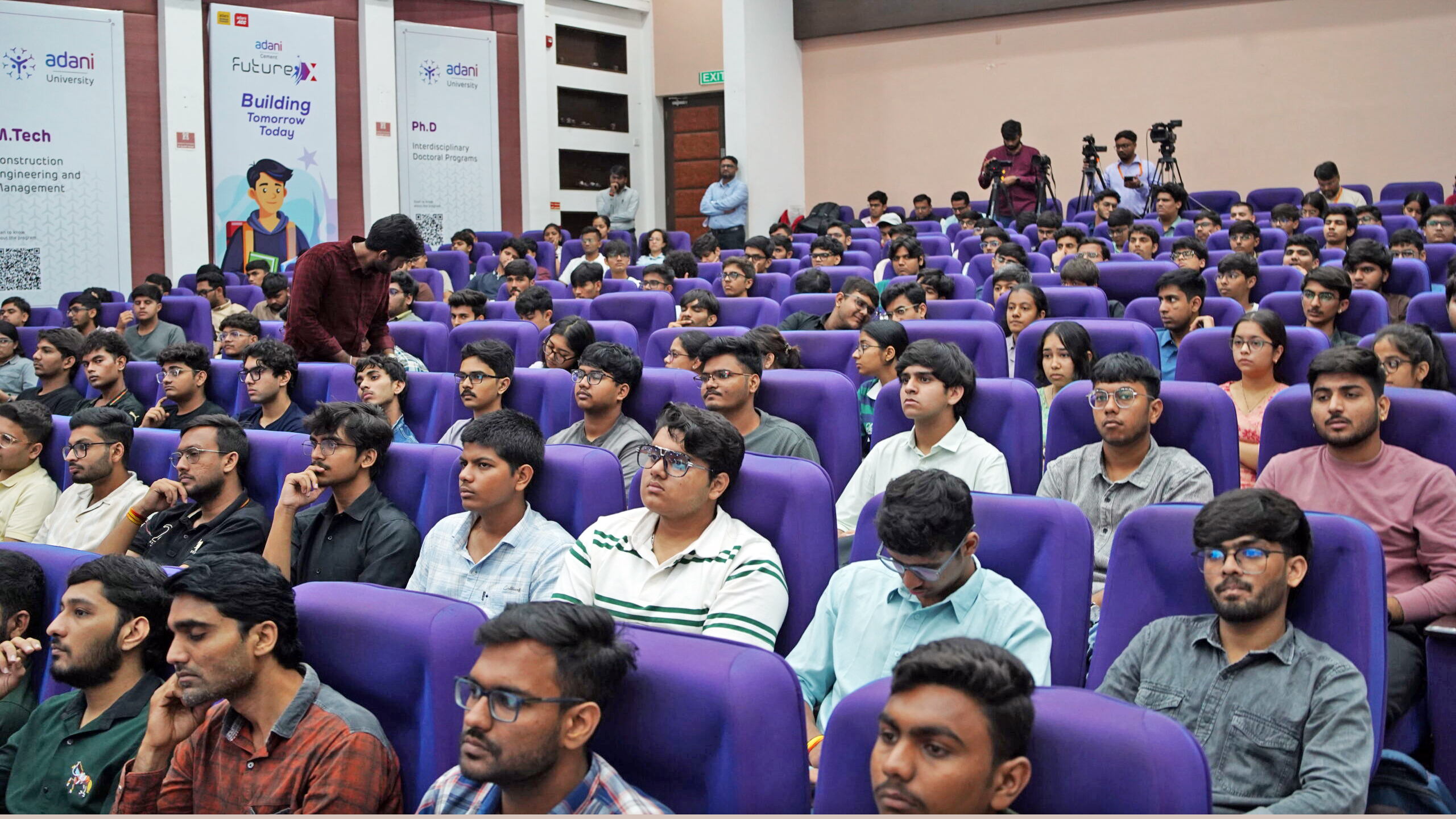 A large group of students are seated in an auditorium, listening to a presentation at Adani University.