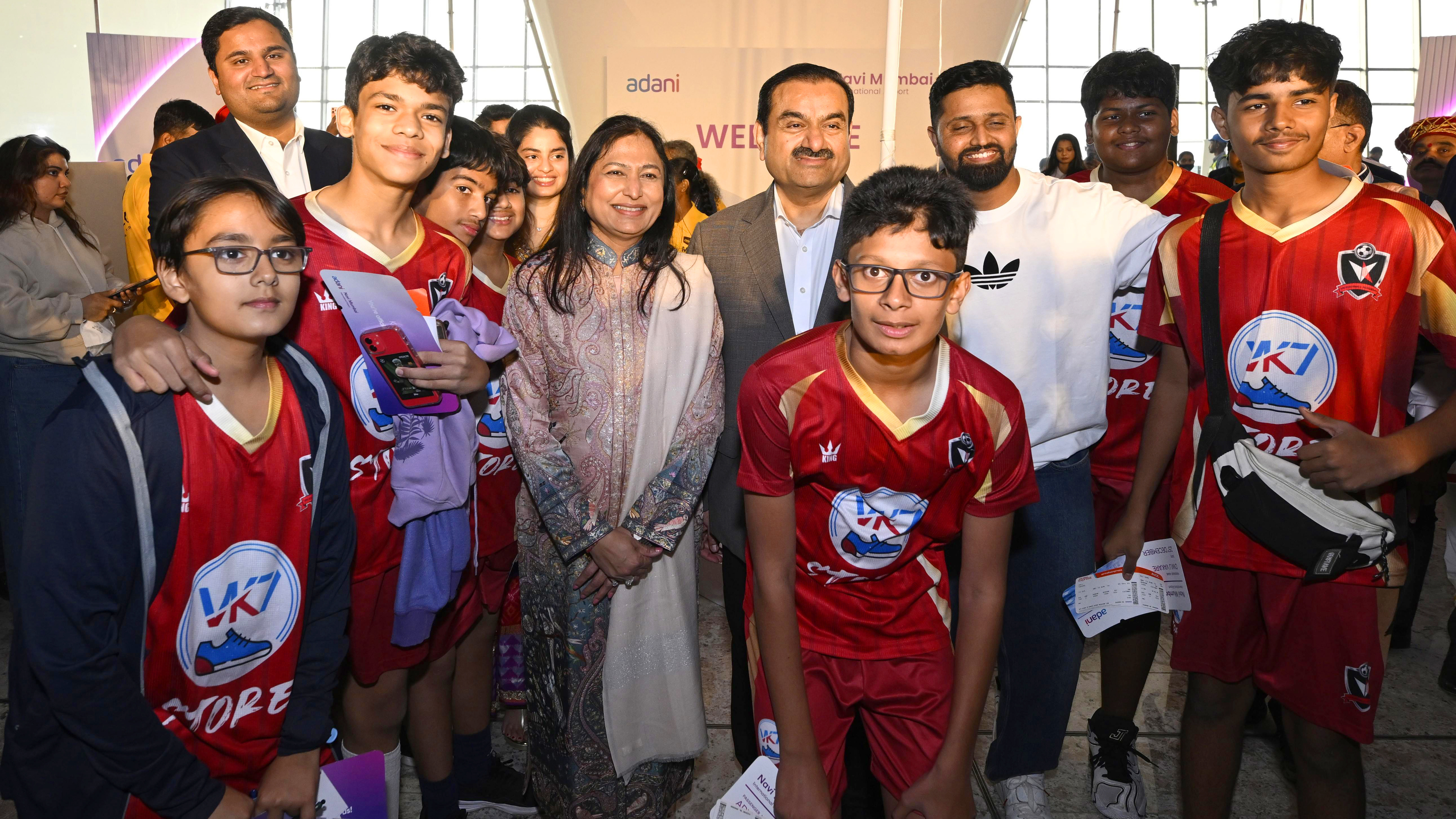 Gautam and Priti Adani pose with a group of young boys in red sports jerseys, adults nearby.