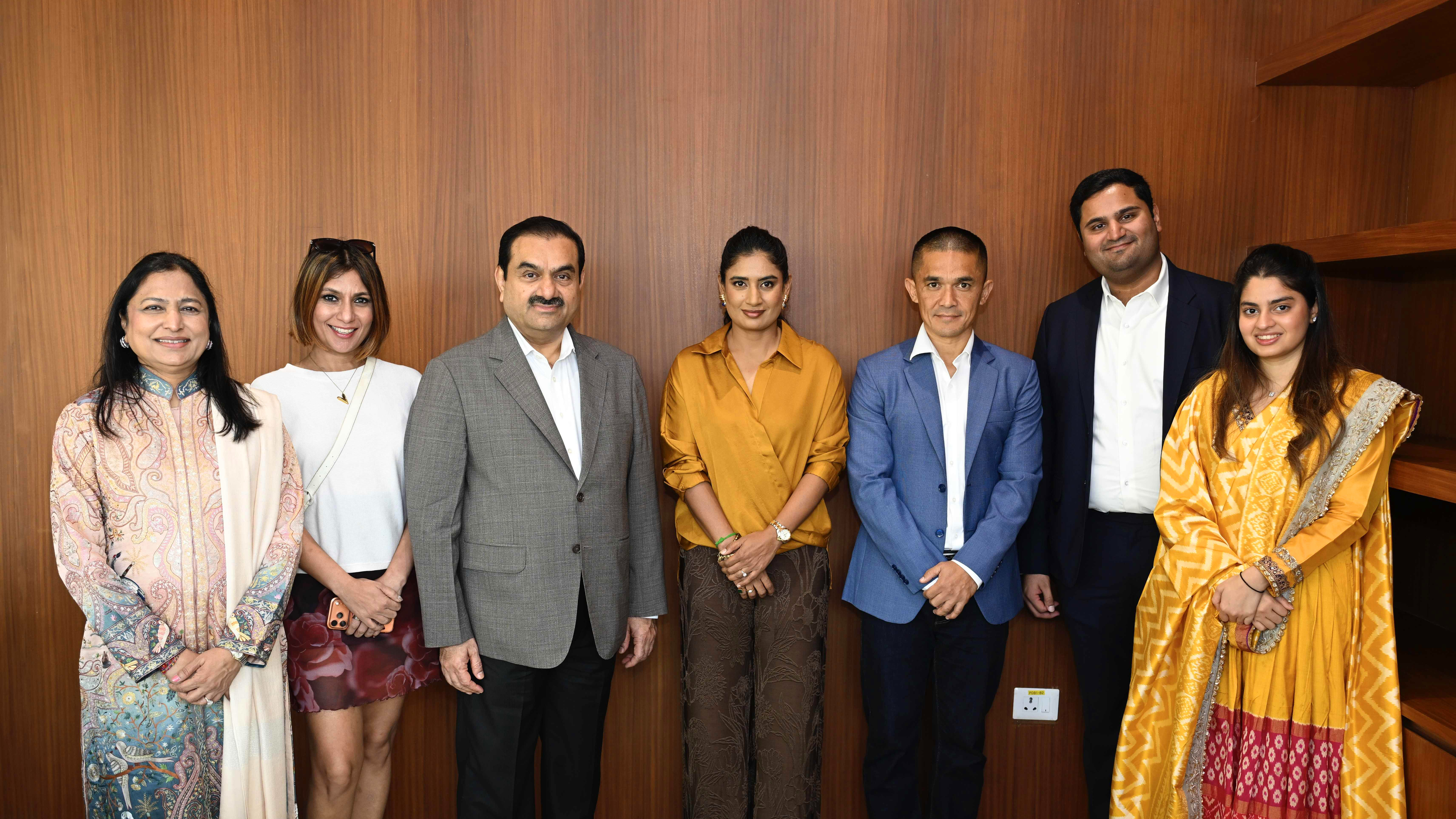 Seven people, including Gautam Adani, stand smiling in front of a wooden wall.