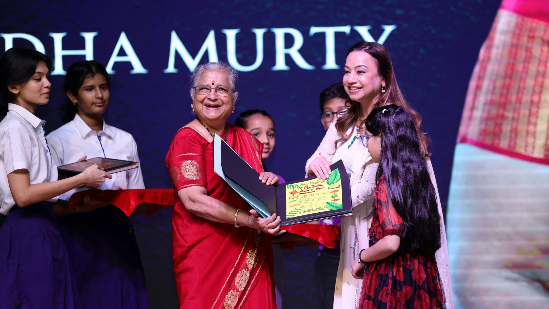 Sudha Murty smiling and receiving a framed gift from a woman and child, with students on stage.
