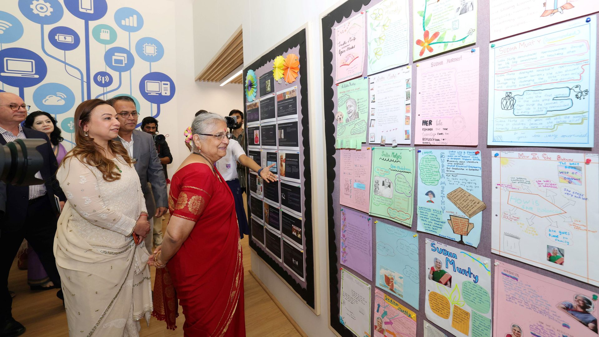Sudha Murty and others view student projects about her on a wall display.