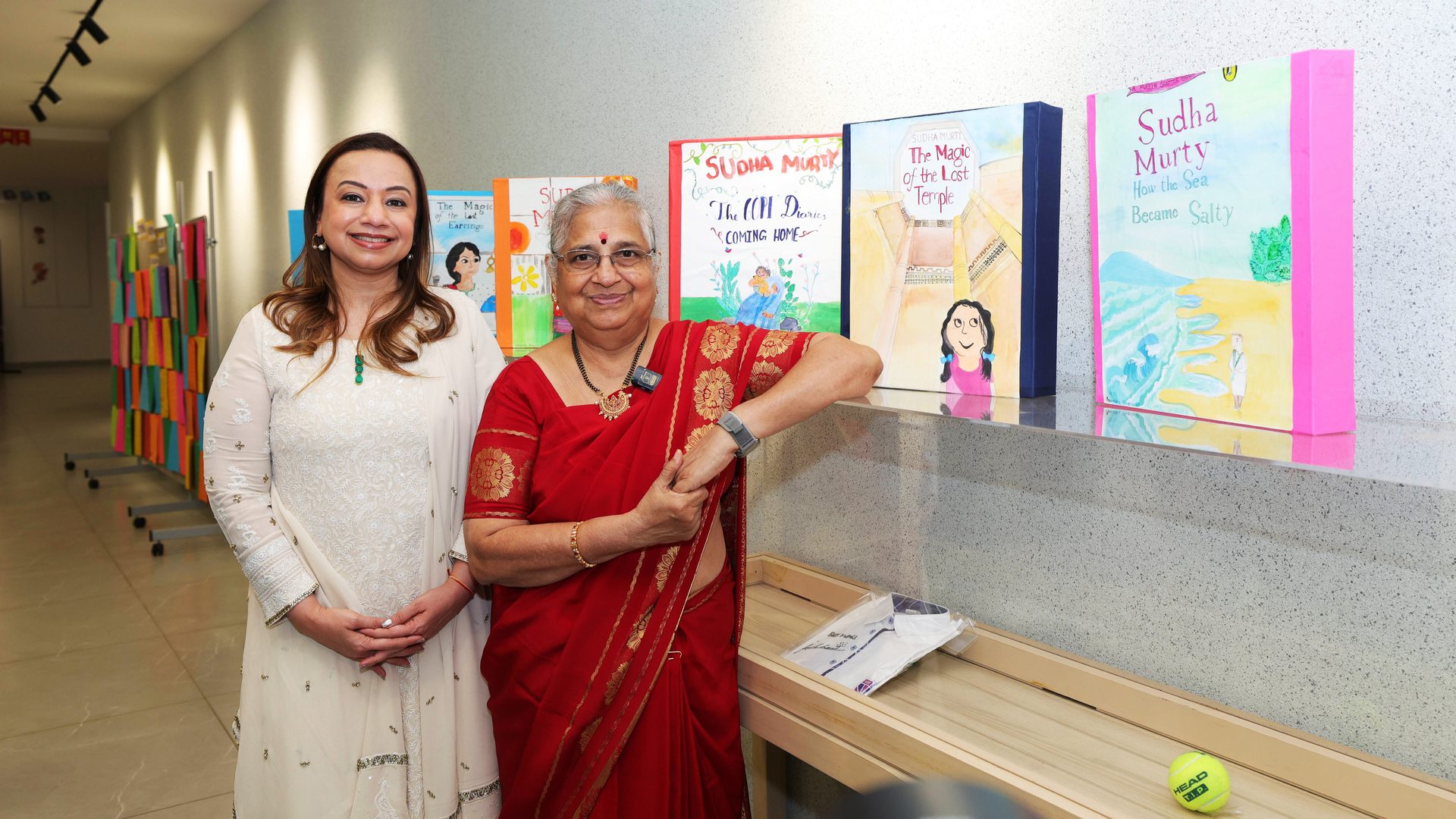 Sudha Murty and another woman smile beside large displays of Murty's books.