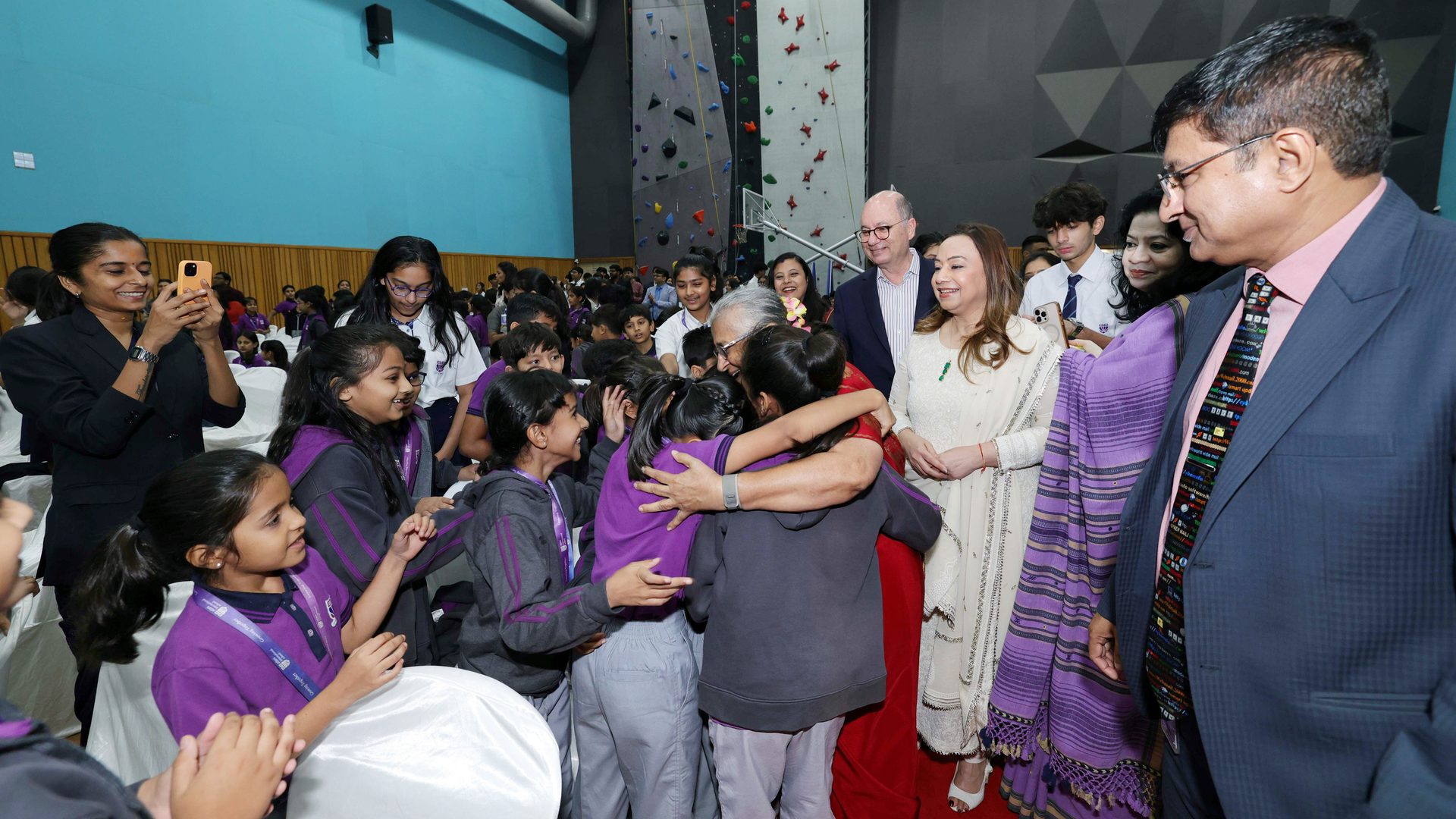 An older woman warmly hugs schoolgirls in uniform, surrounded by smiling adults and students at an indoor event.