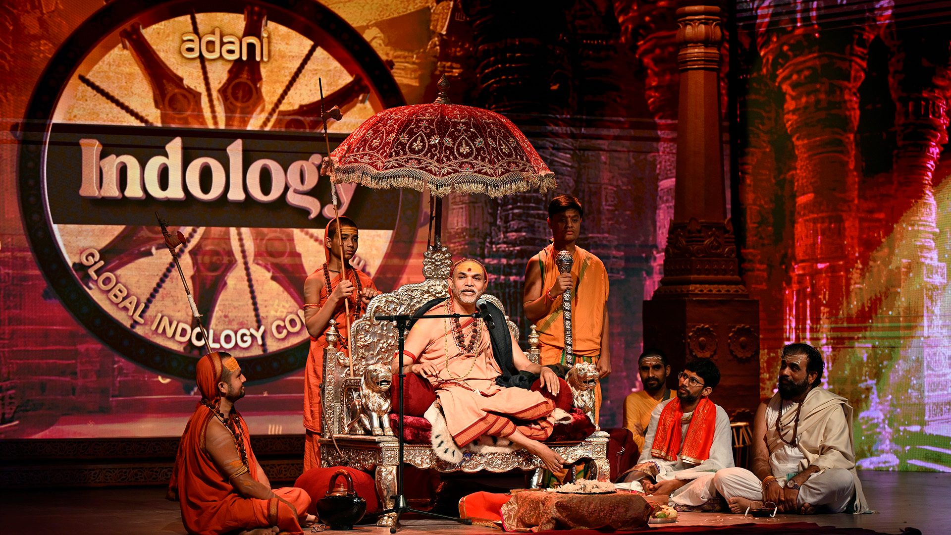 Spiritual leader on ornate throne with followers and 'Adani Indology' backdrop at a cultural event.