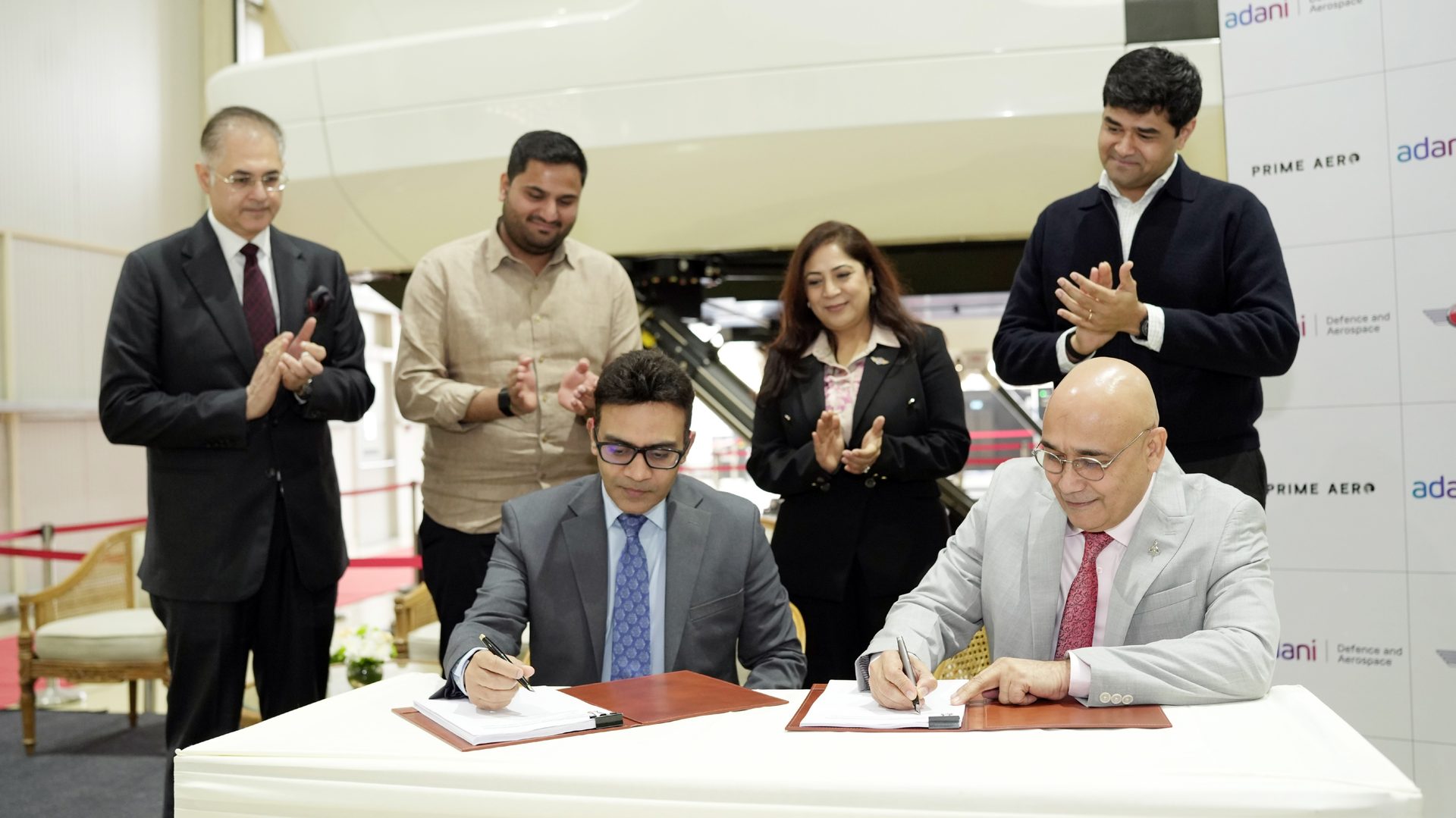 Two men signing documents at a table, while four others clap behind them at an aerospace event.