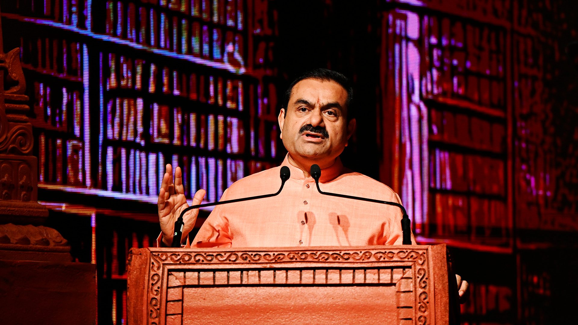 Gautam Adani speaking at a podium with a vibrant digital bookshelf background.