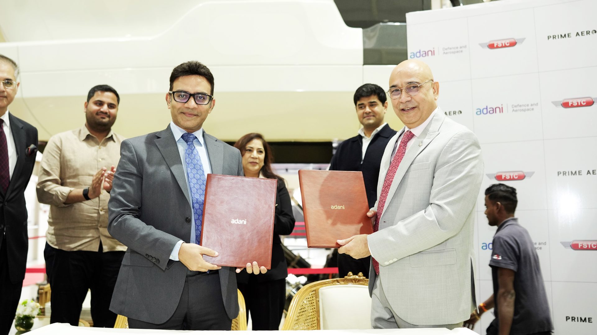 Two men in suits exchange branded documents at a business event, with Adani logos visible.