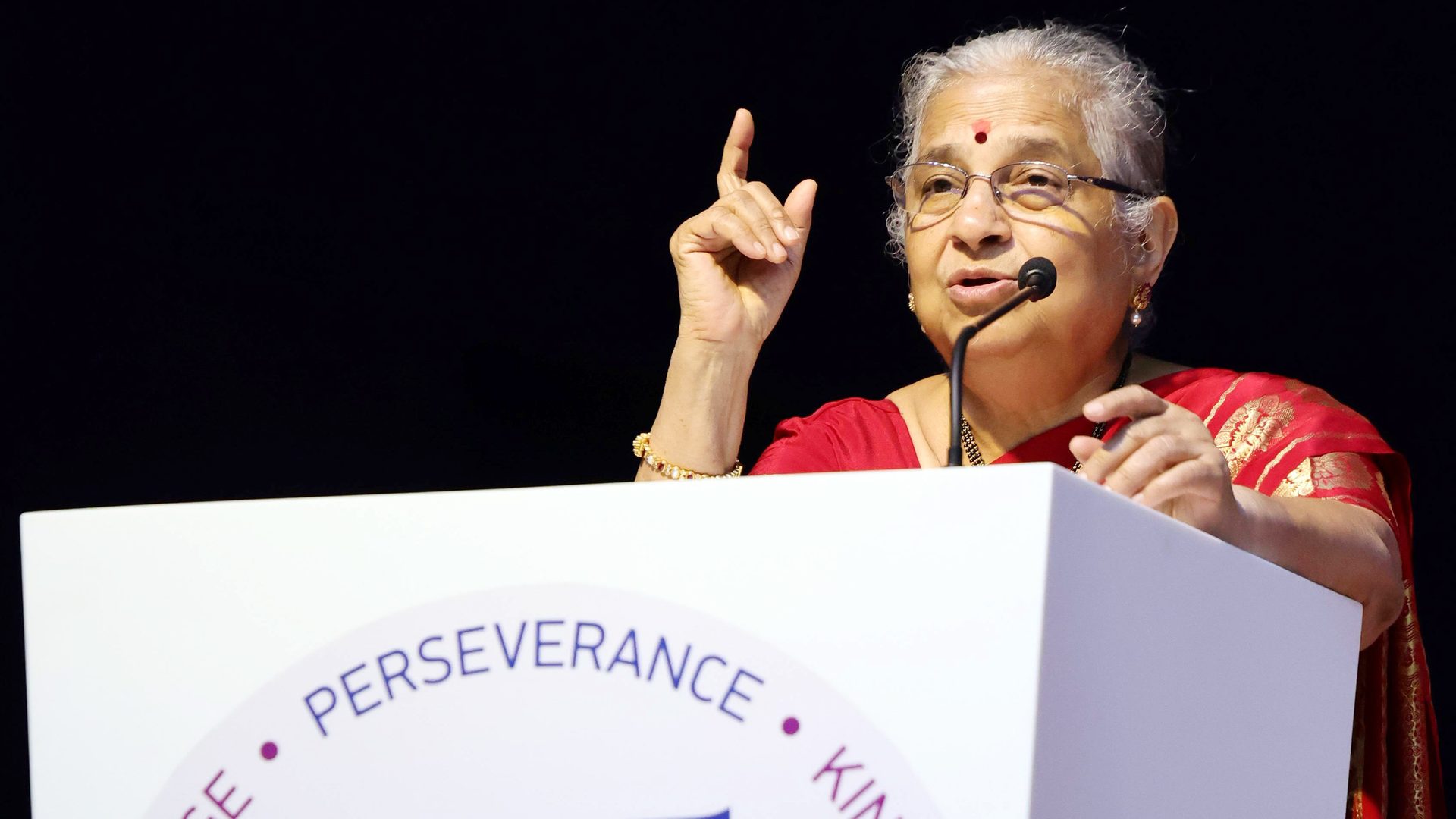 Sudha Murty, in a red saree, speaking at a podium with a raised hand.