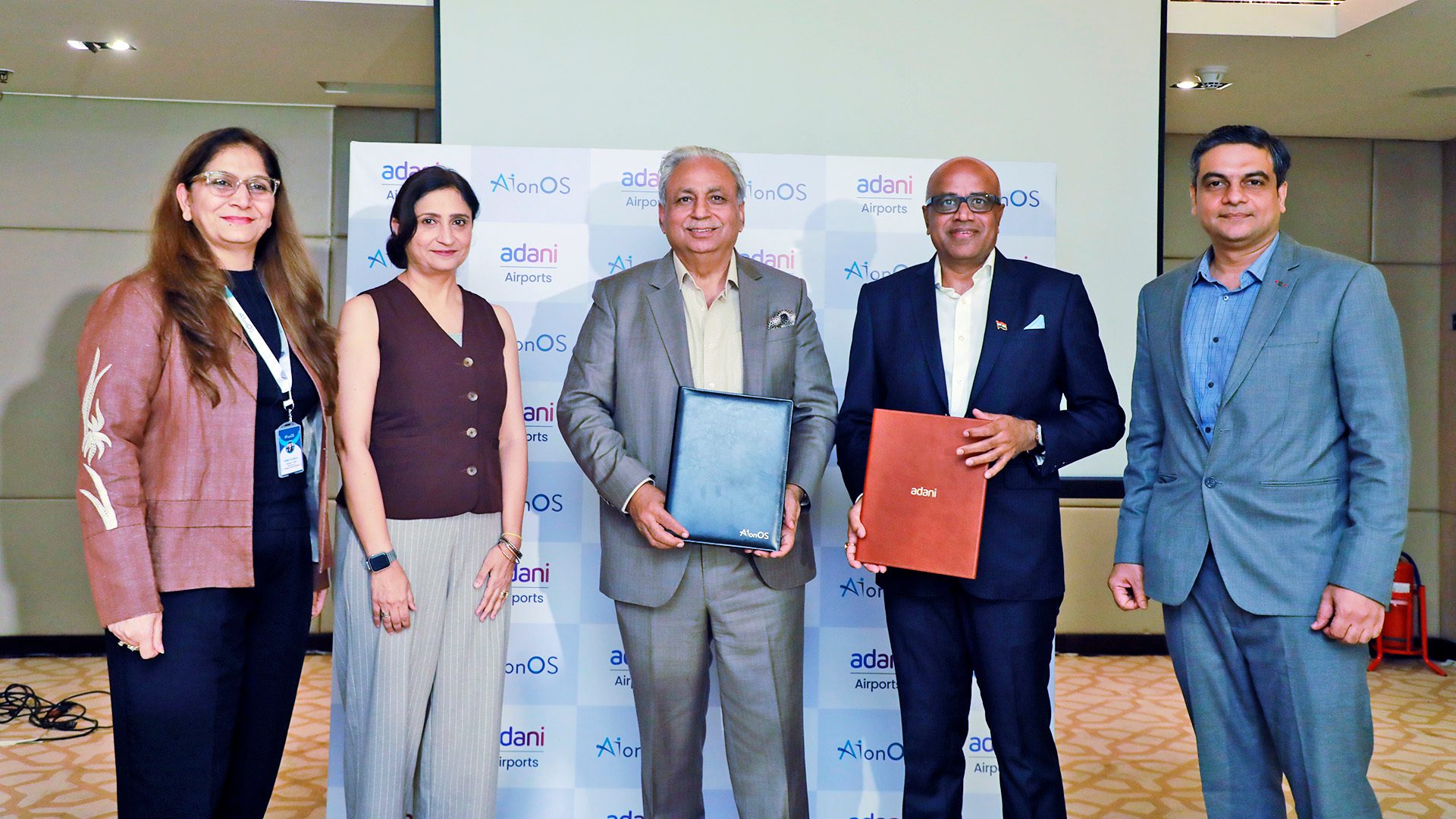 Five people, three men holding folders, stand before Adani Airports and AionOS logos, indicating a partnership.