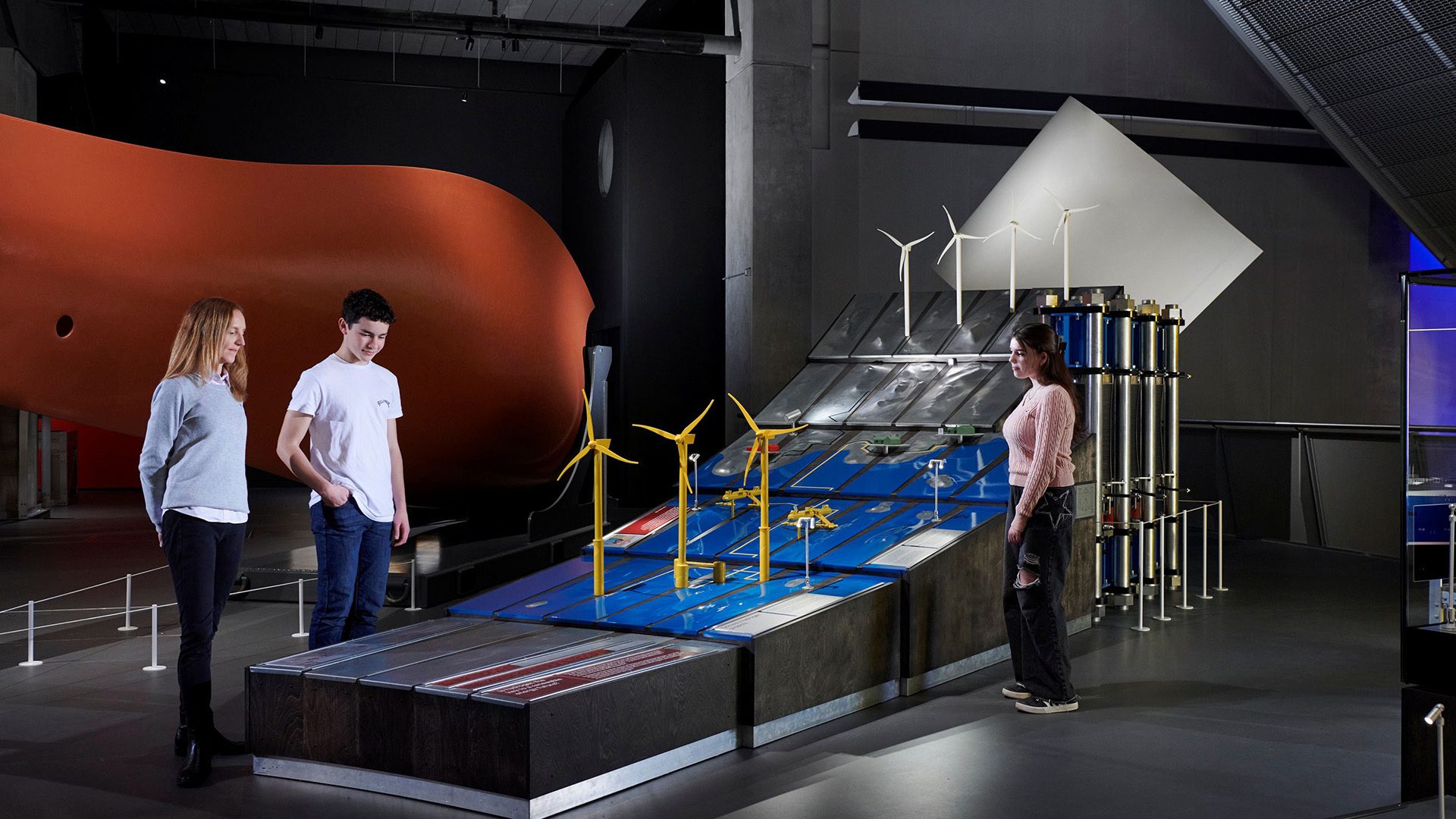 Three people examine wind turbine models and a large orange hull in a museum exhibition.