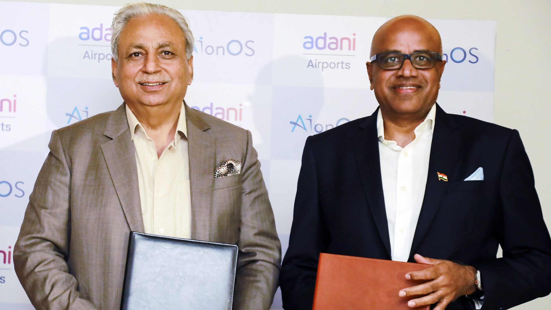 Two smiling men hold documents; Adani Airports and AionOS logos in the background.