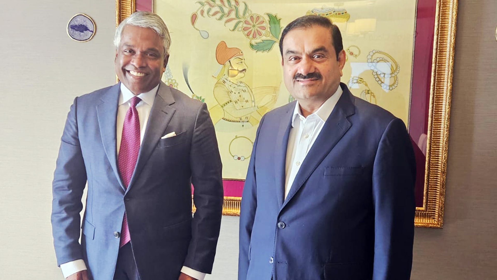 Two men in suits, Gautam Adani and P. Sukumar, smile in front of traditional Indian artwork.