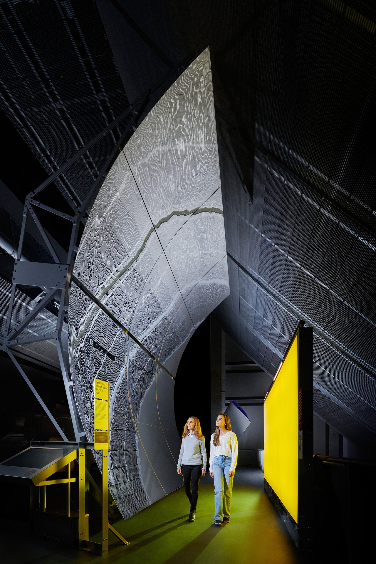Two women walk past a large, curved patterned screen and a yellow lit wall in a dim exhibit.