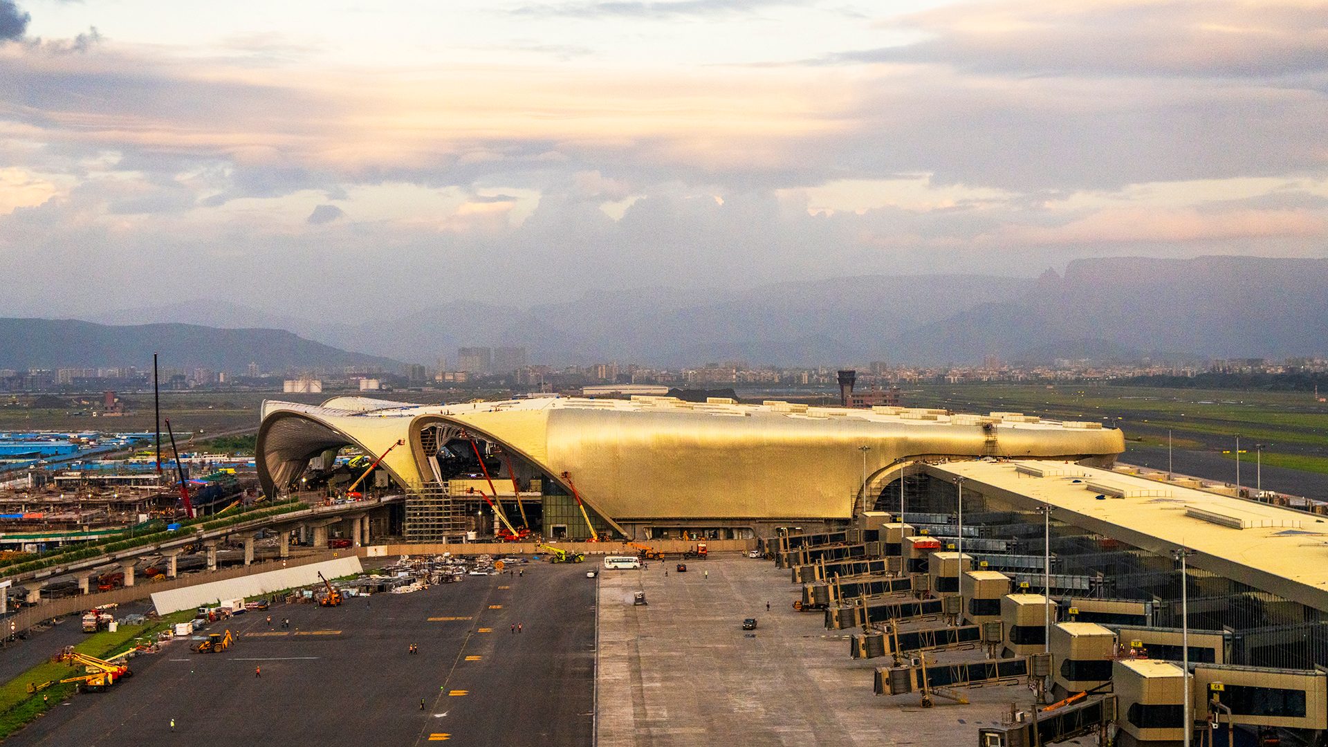 Golden airport terminal under construction, with mountains and city in the background.