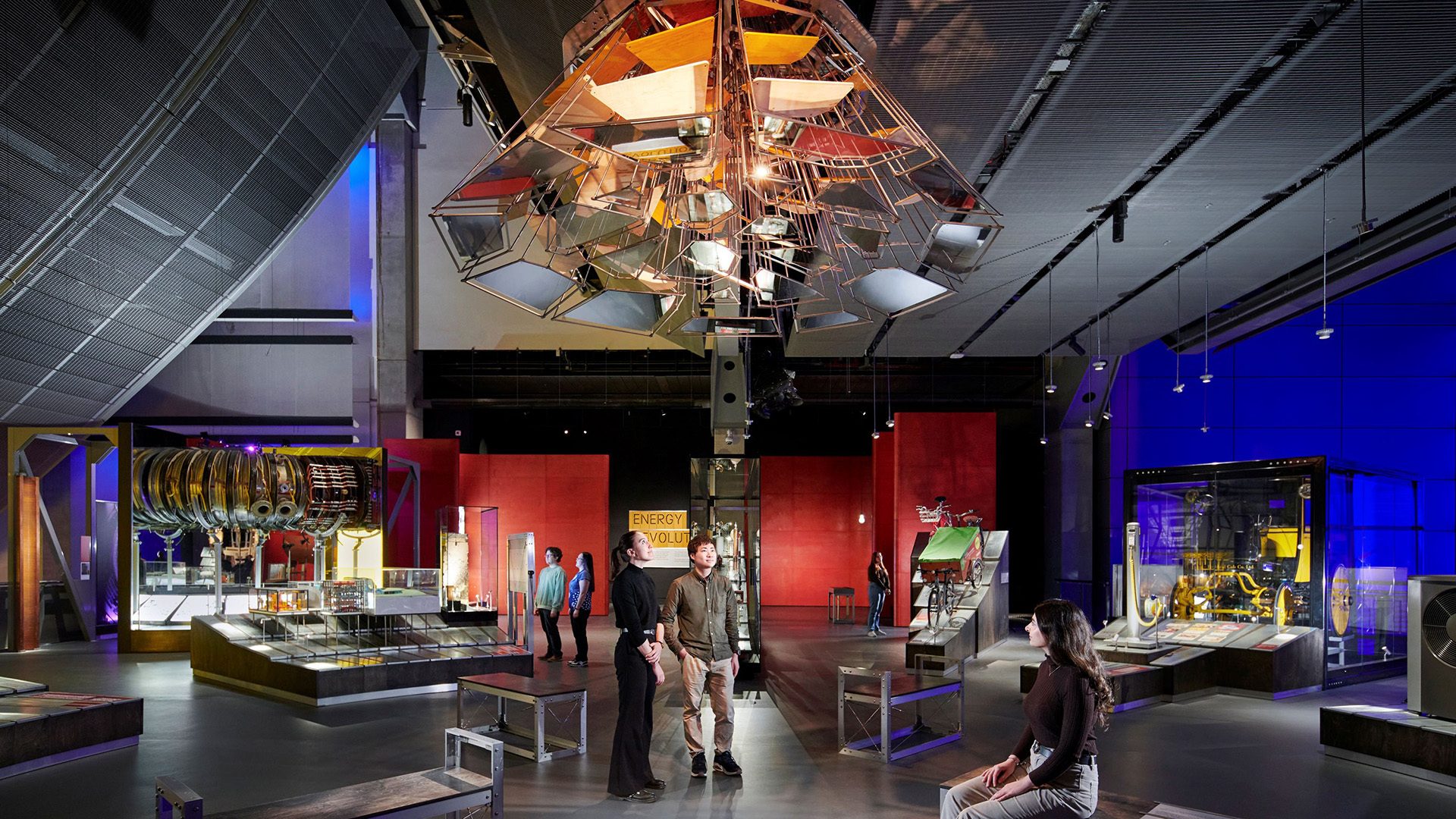 People explore a museum exhibit featuring a geometric chandelier and large mechanical displays.