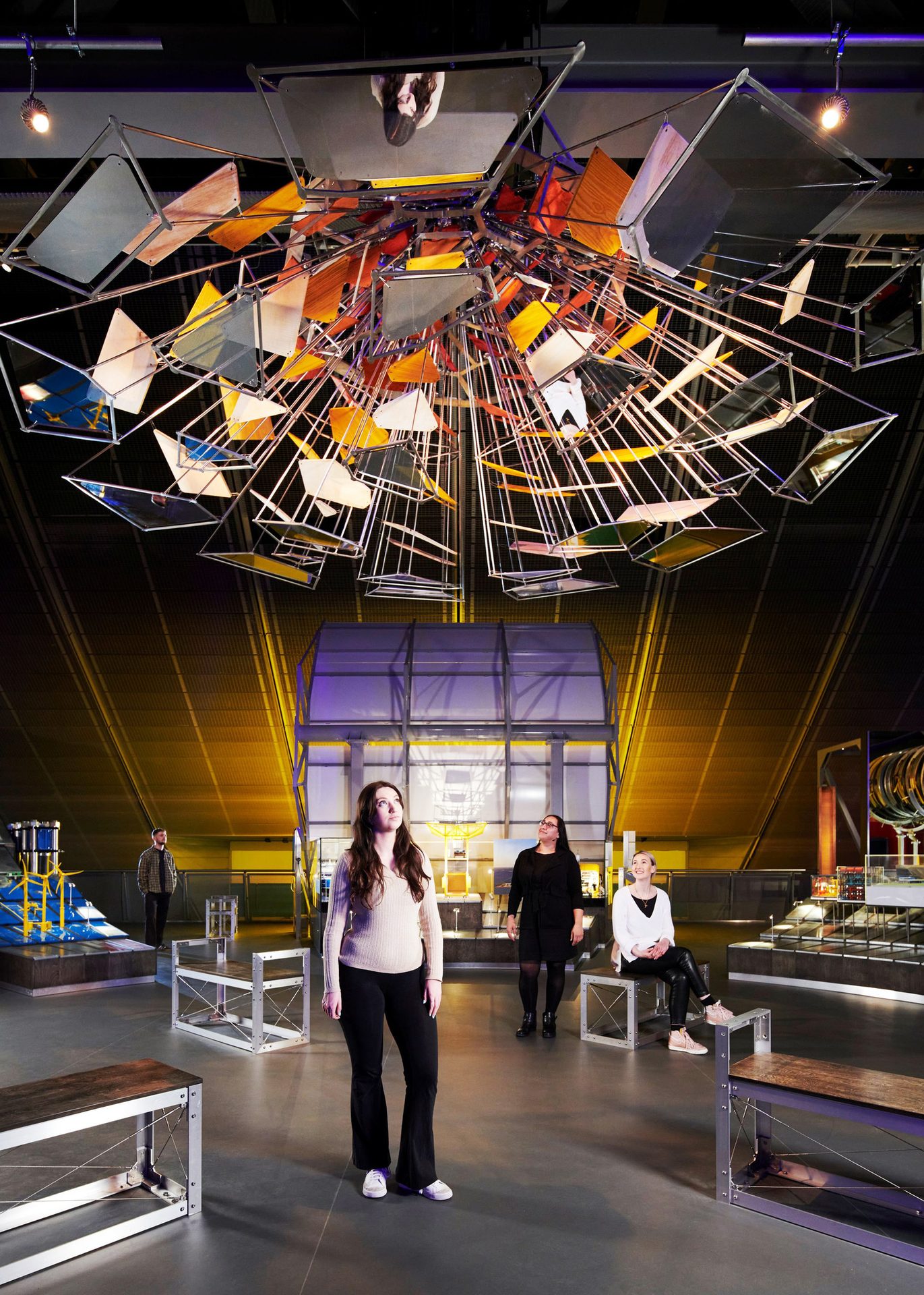 People admiring a large, colorful, geometric mobile art installation in a spacious hall.