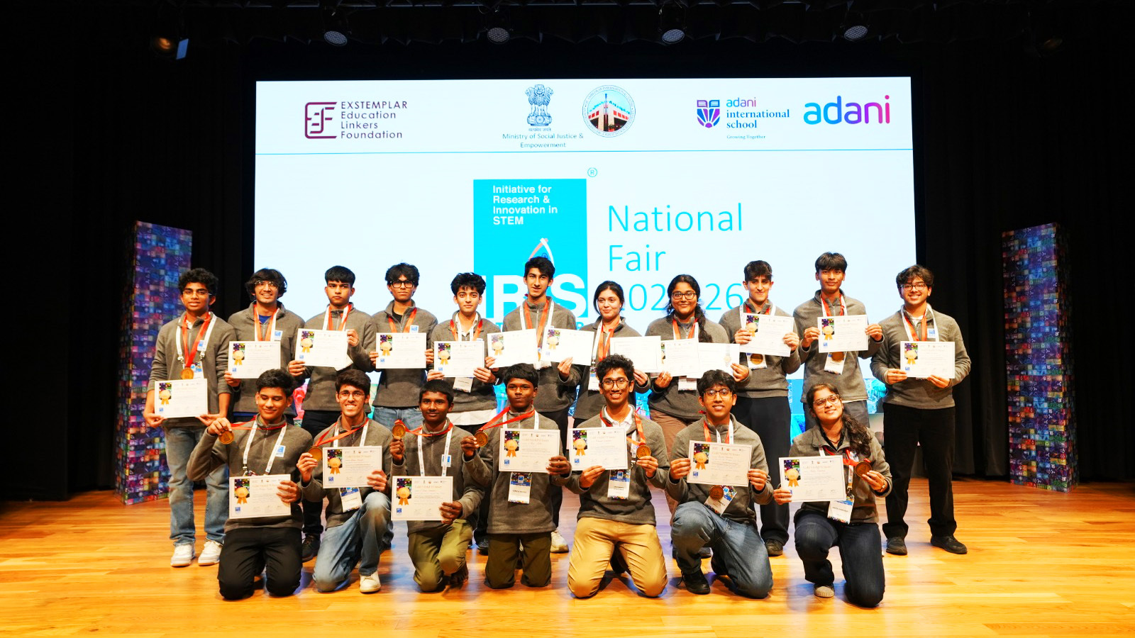 A group of students holding certificates and medals on a stage, with a large screen displaying "National Fair".