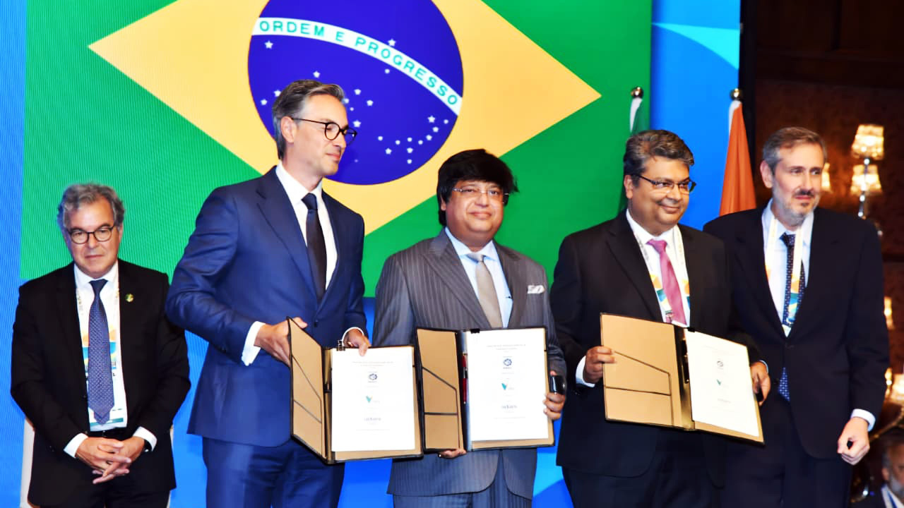 Five men in suits hold documents at a signing ceremony with the Brazilian flag backdrop.
