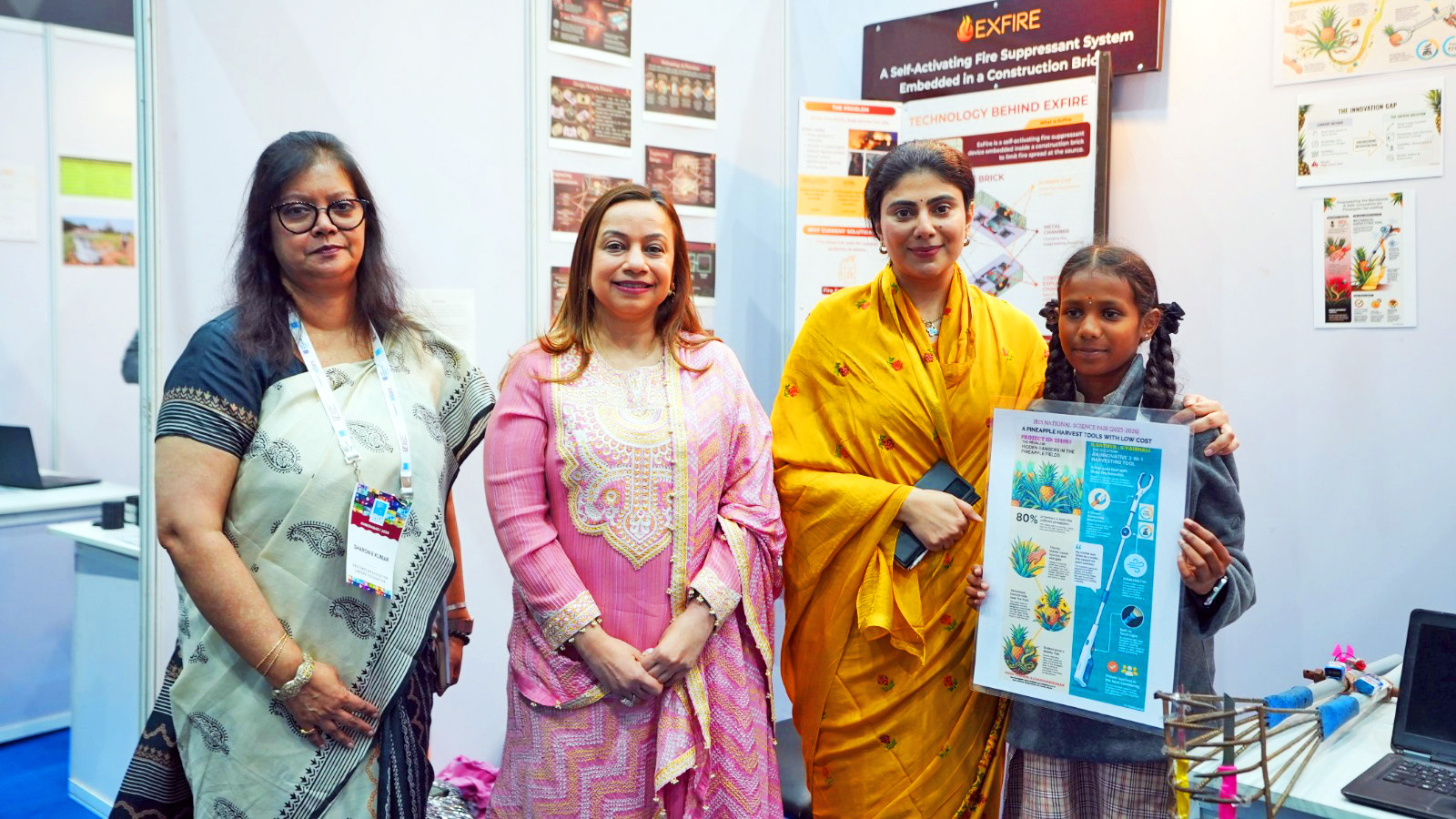 Four females, including a girl holding a poster, stand together at an exhibition booth.