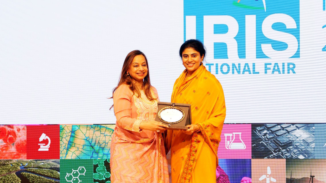 Two women, one in a peach outfit and one in a yellow saree, holding a plaque at an event.