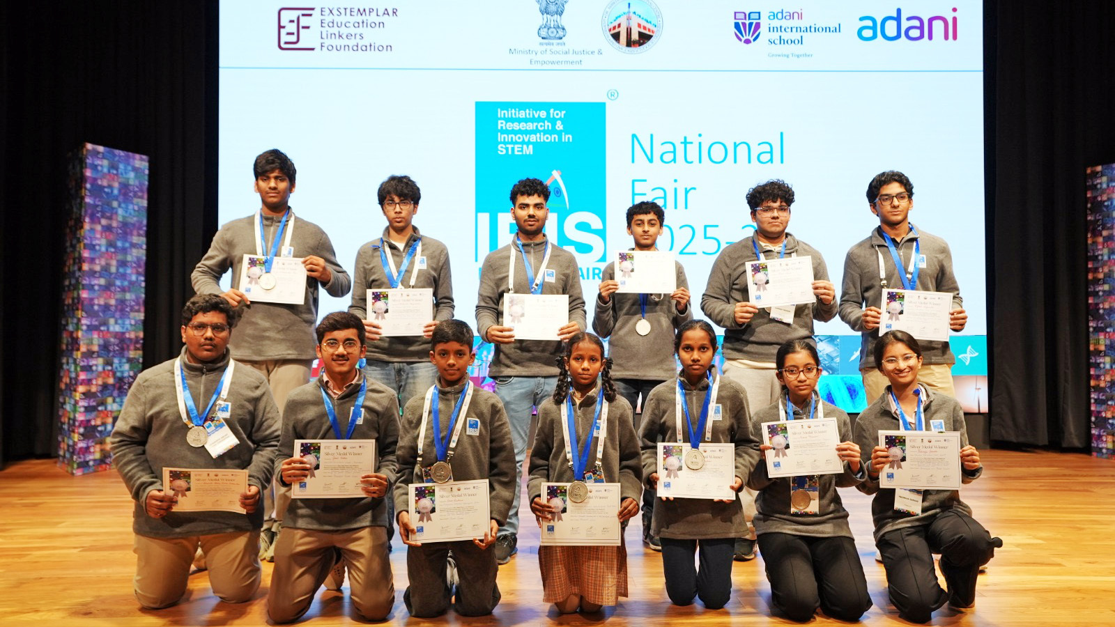 Group of students proudly displaying certificates and medals on a stage at a STEM fair.