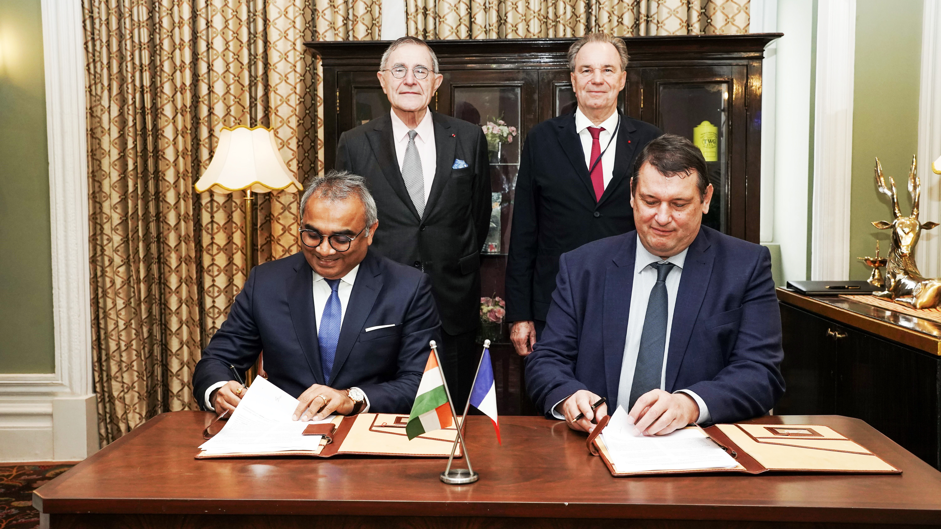 Four men in suits, two signing documents at a table with Indian and French flags, two standing behind them.