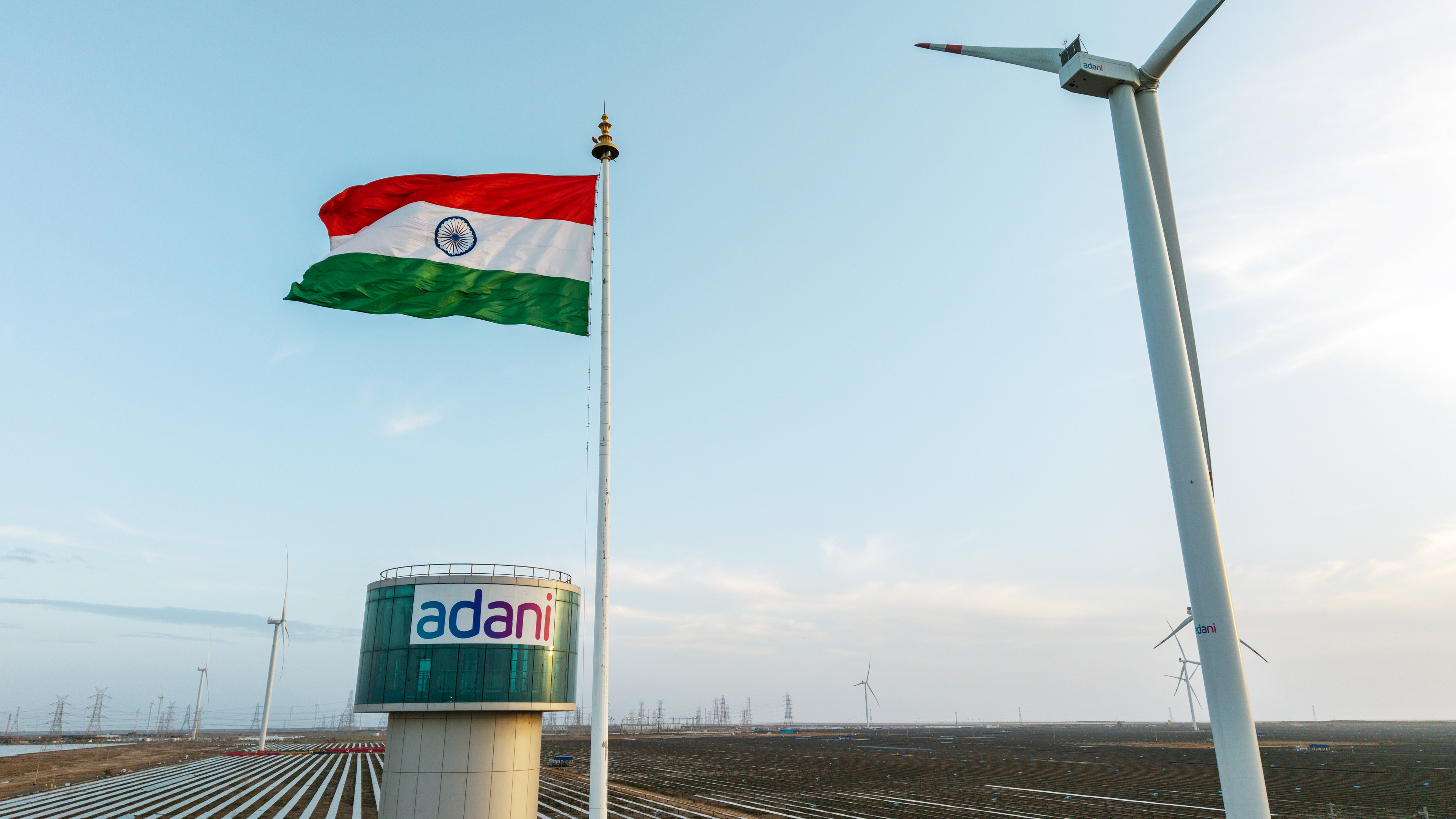 Indian flag, Adani logo, wind turbines, and solar panels against a blue sky, representing renewable energy.