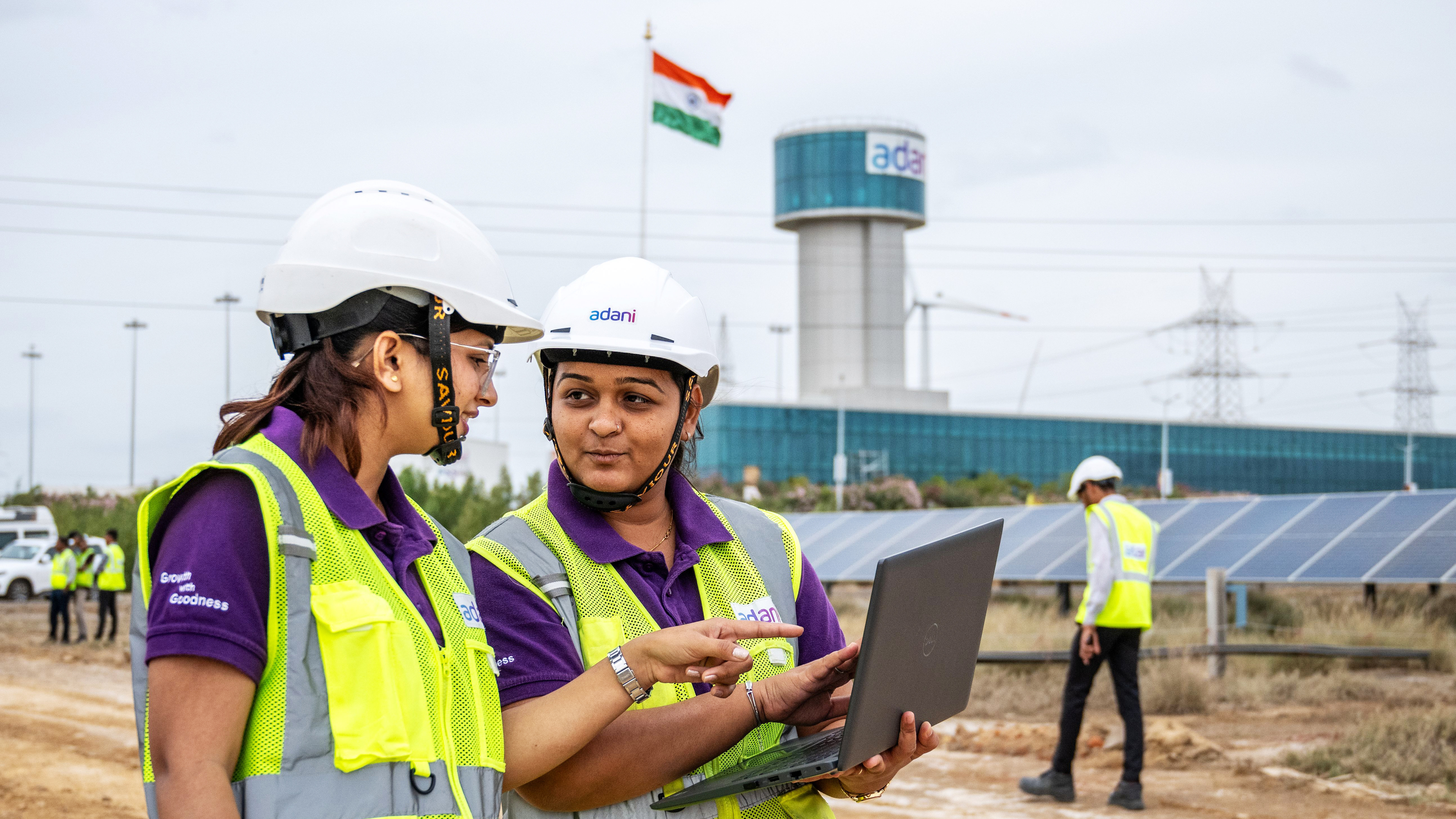 Two women engineers in hard hats and vests discuss plans on a laptop at a solar plant with an Indian flag and Adani tower.