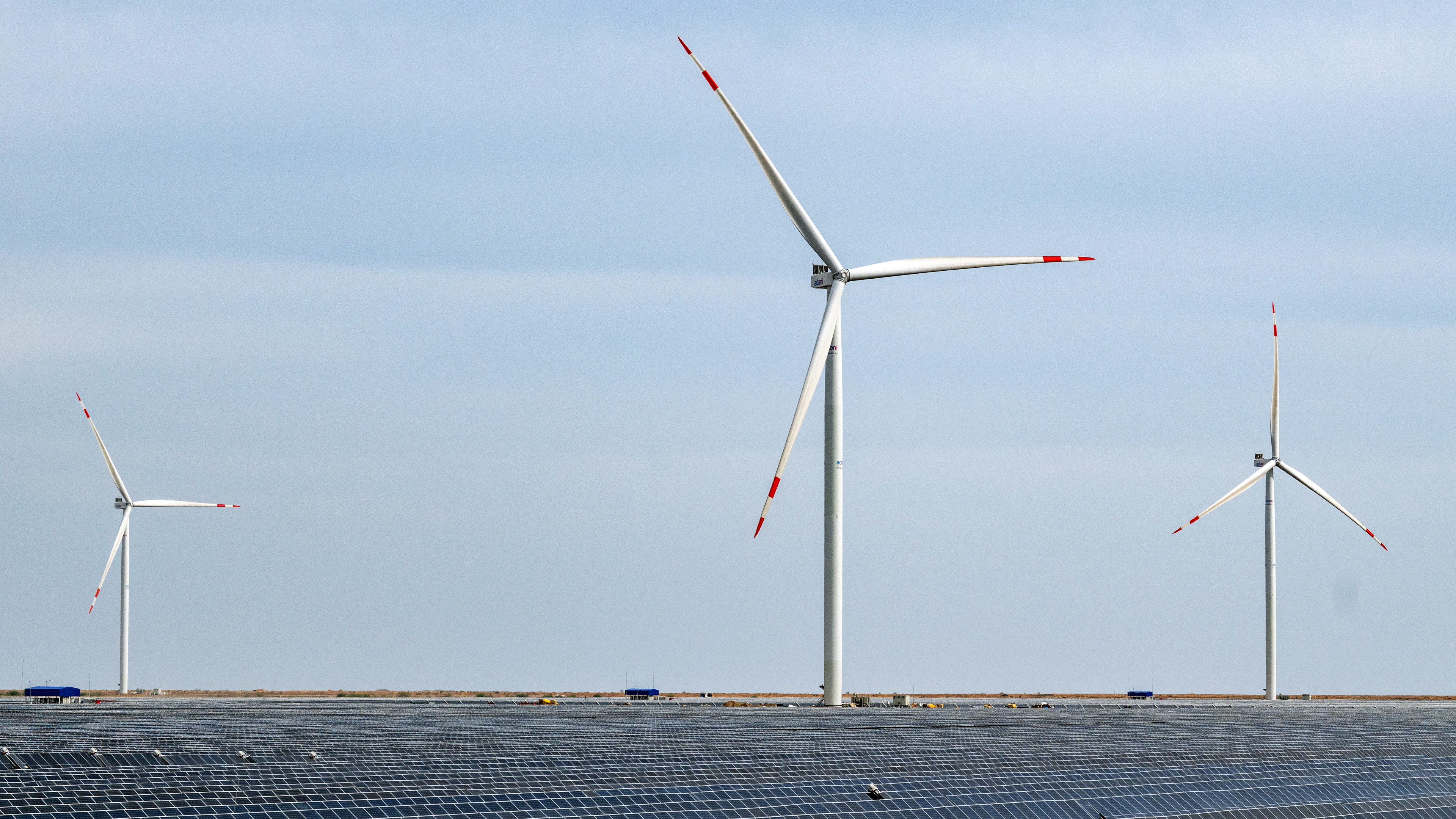 Three wind turbines stand over a field of solar panels under a clear sky, showing renewable energy.