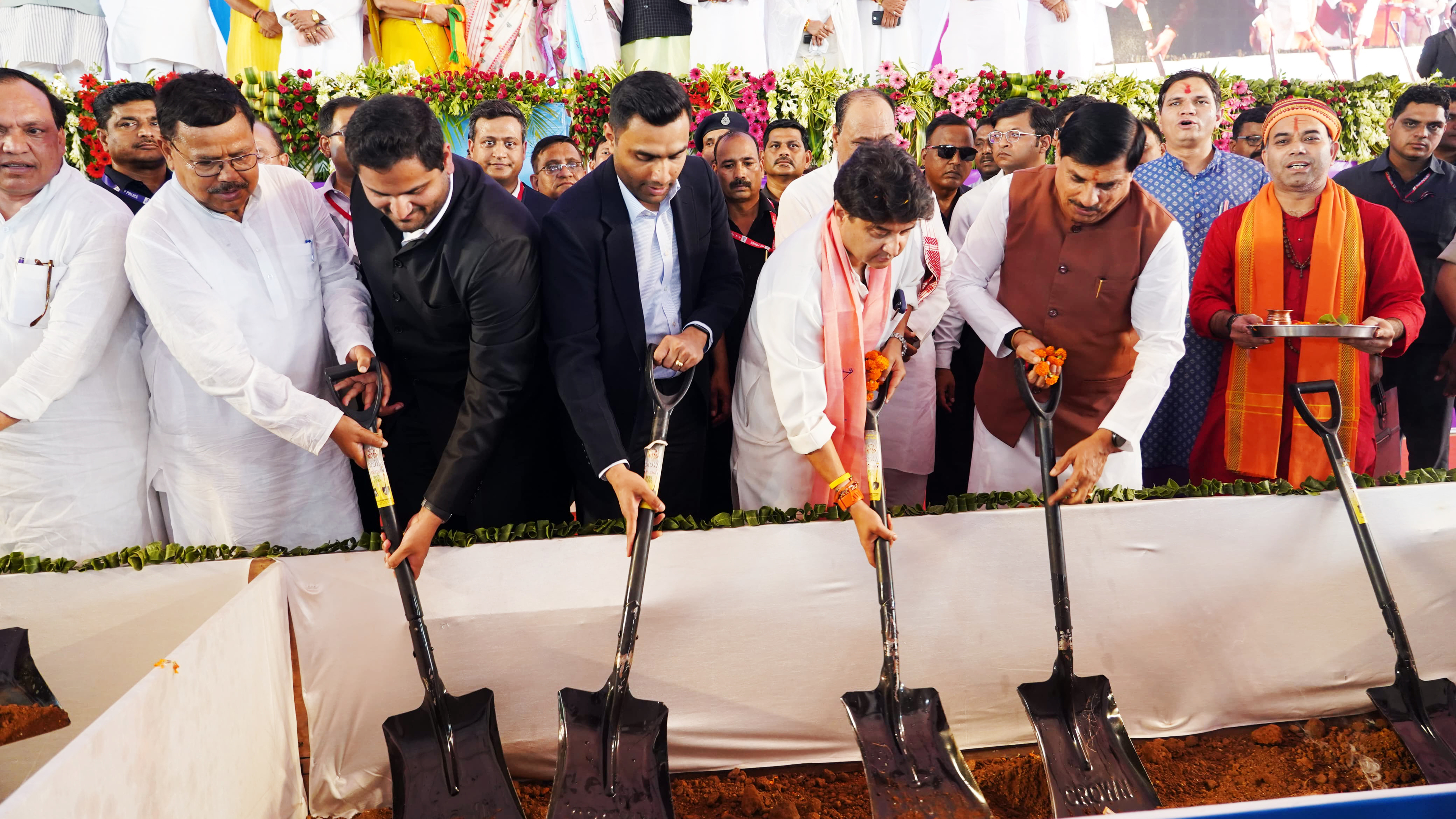 A group of men, mostly in traditional Indian attire, participate in a groundbreaking ceremony with shovels.