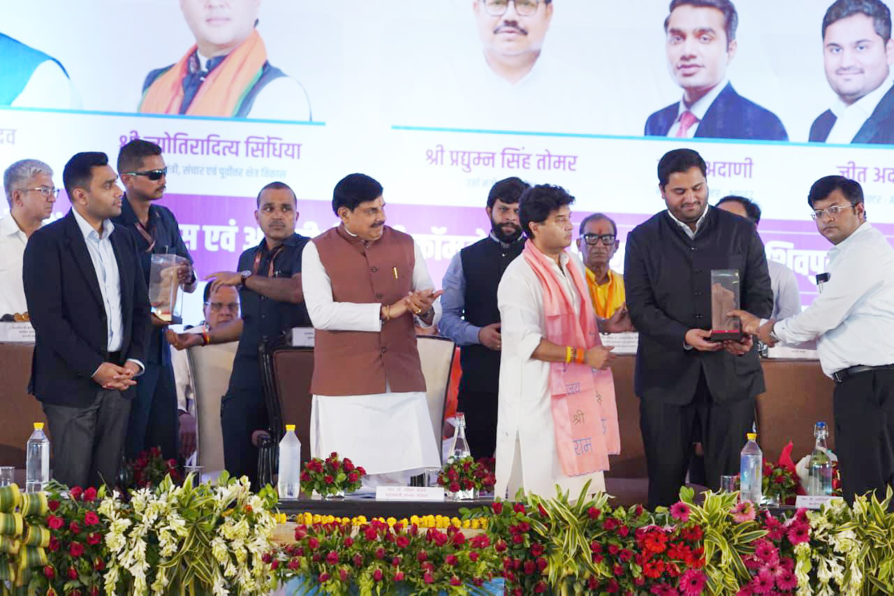 Men on stage at an event; one man in a black jacket receives an award, while others applaud and observe.