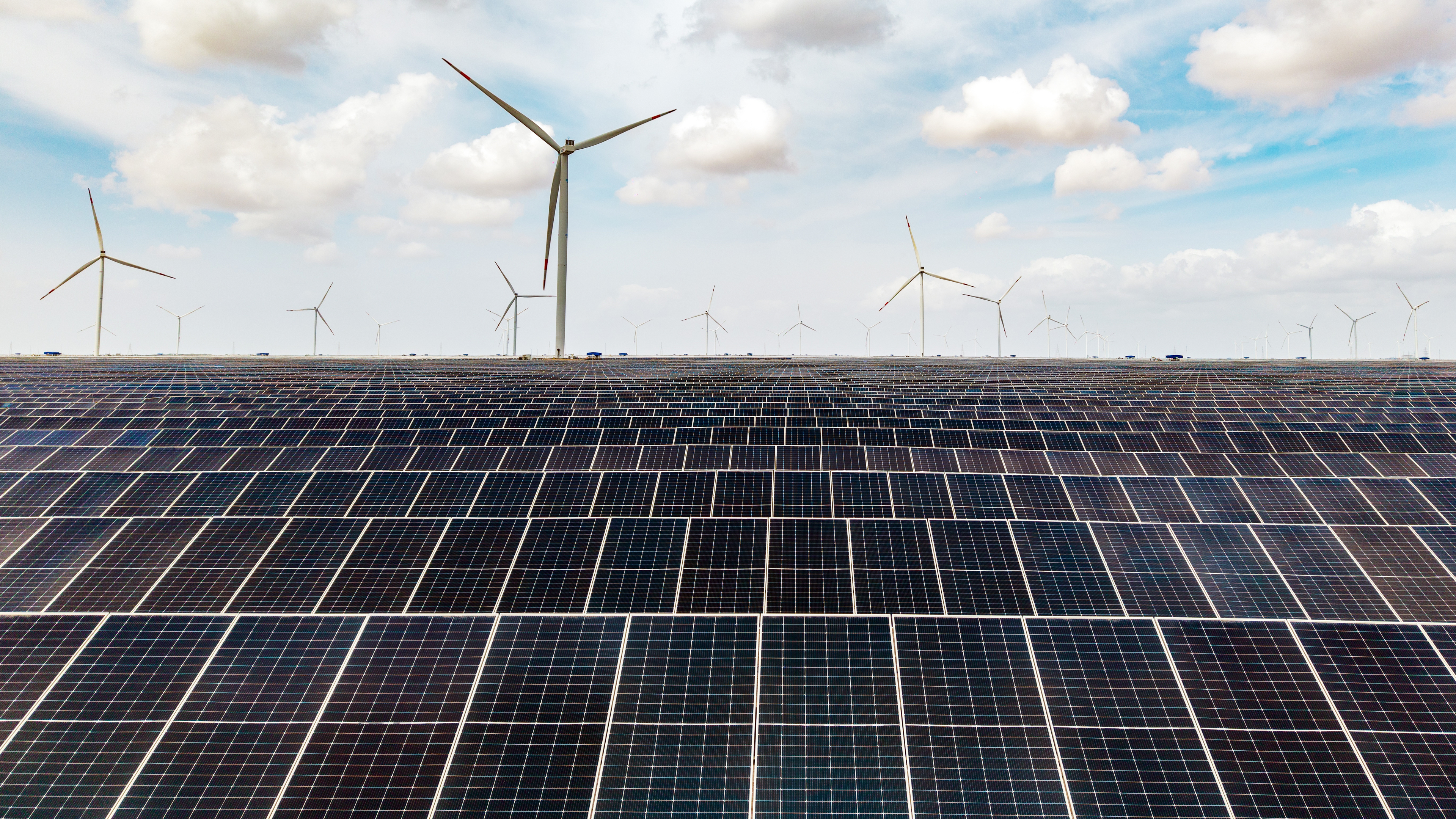 Vast solar panels fill the foreground with many wind turbines in the distance, showcasing a renewable energy farm.