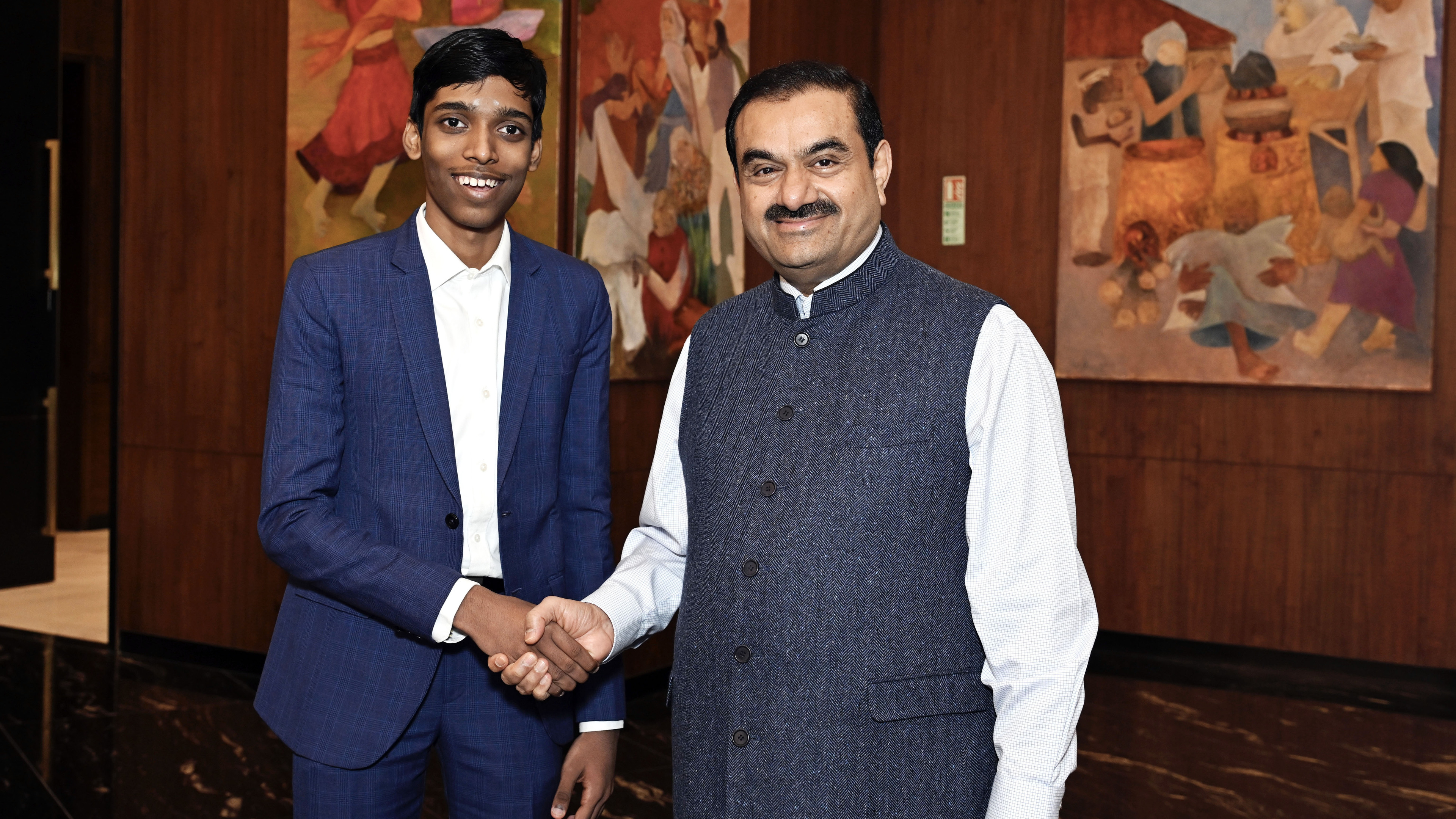 A young man shakes hands with Gautam Adani in a room with wooden walls and colorful paintings.