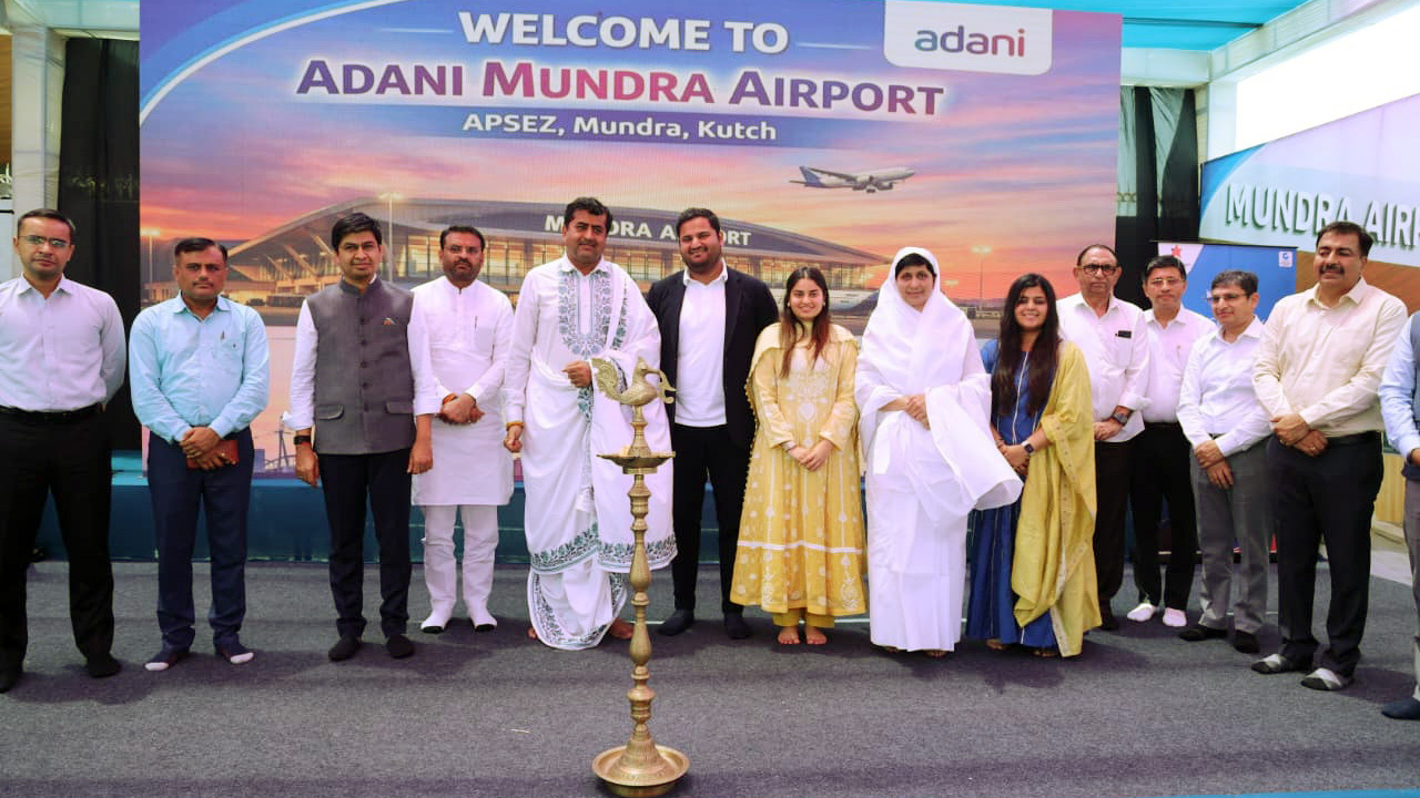 A group of people, including men and women, stand in front of a 'Welcome to Adani Mundra Airport' banner with a traditional lamp in the foreground.