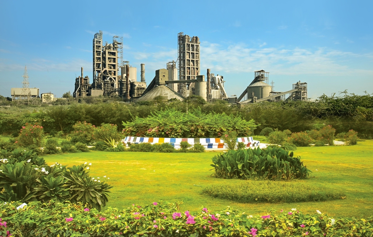A large industrial cement plant in the background, with a vibrant green landscaped garden in the foreground.