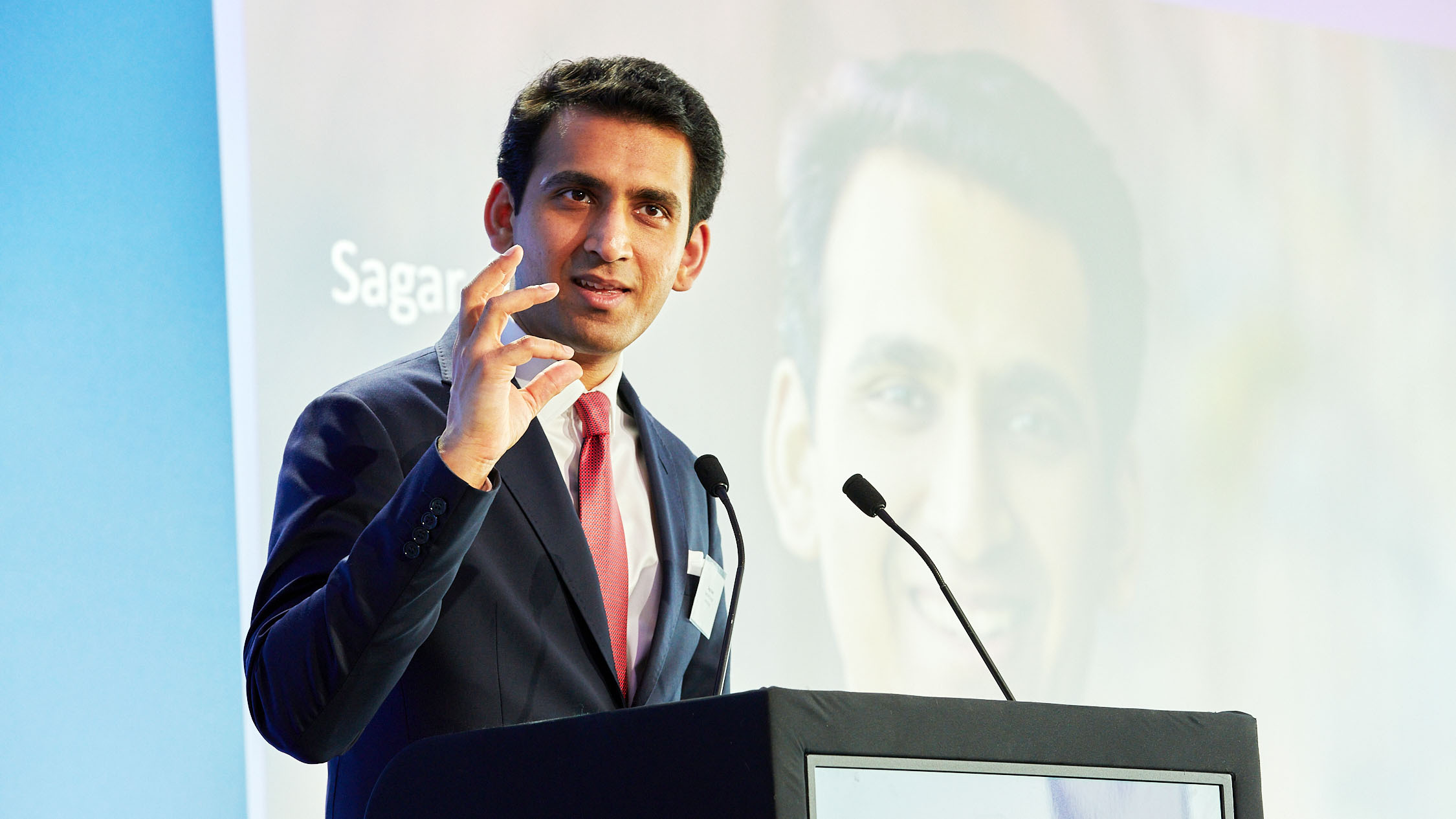 A man in a suit speaks at a podium, gesturing with his hand.