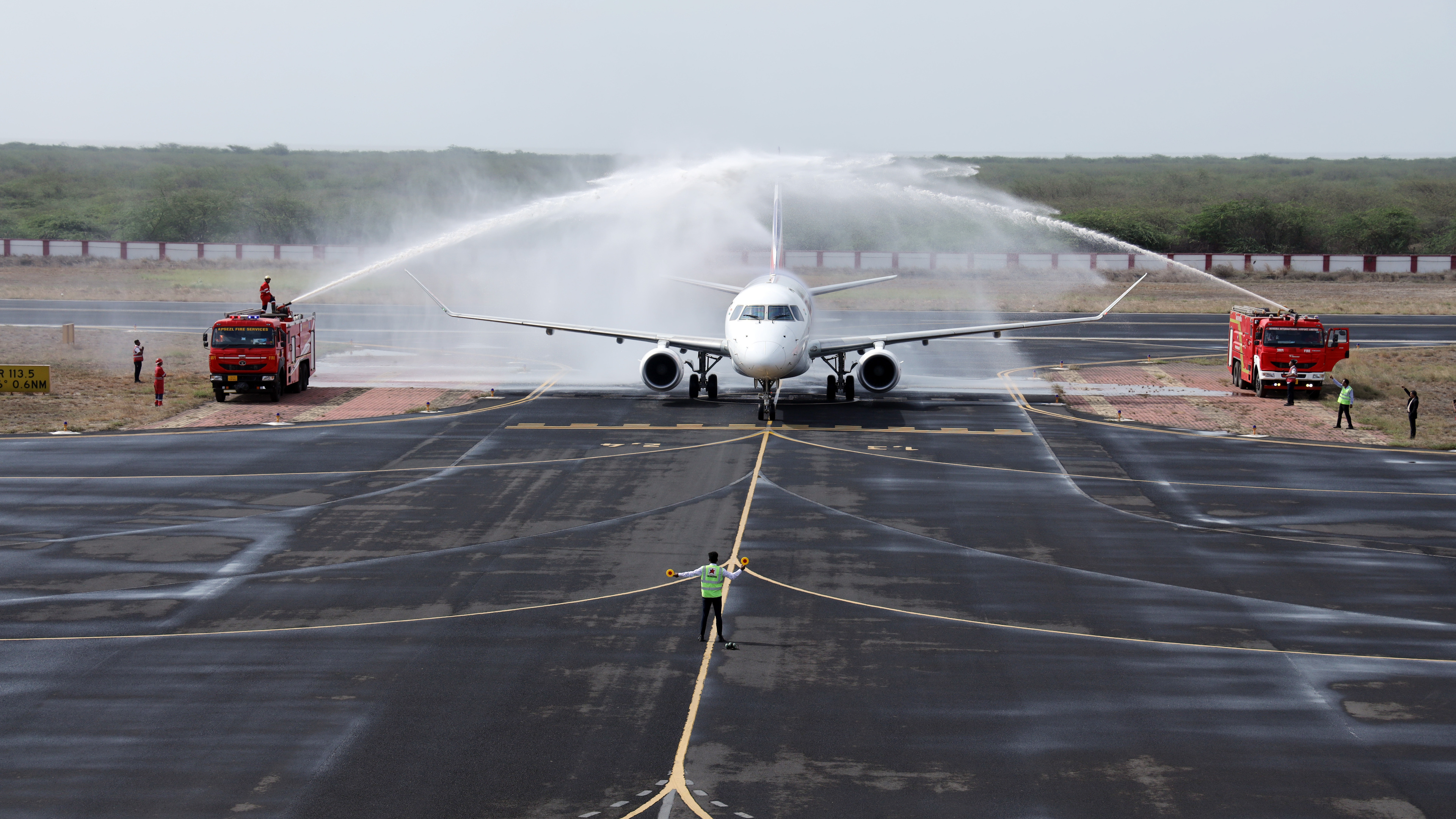 An airplane receives a water salute from two fire trucks on a runway, directed by ground crew.