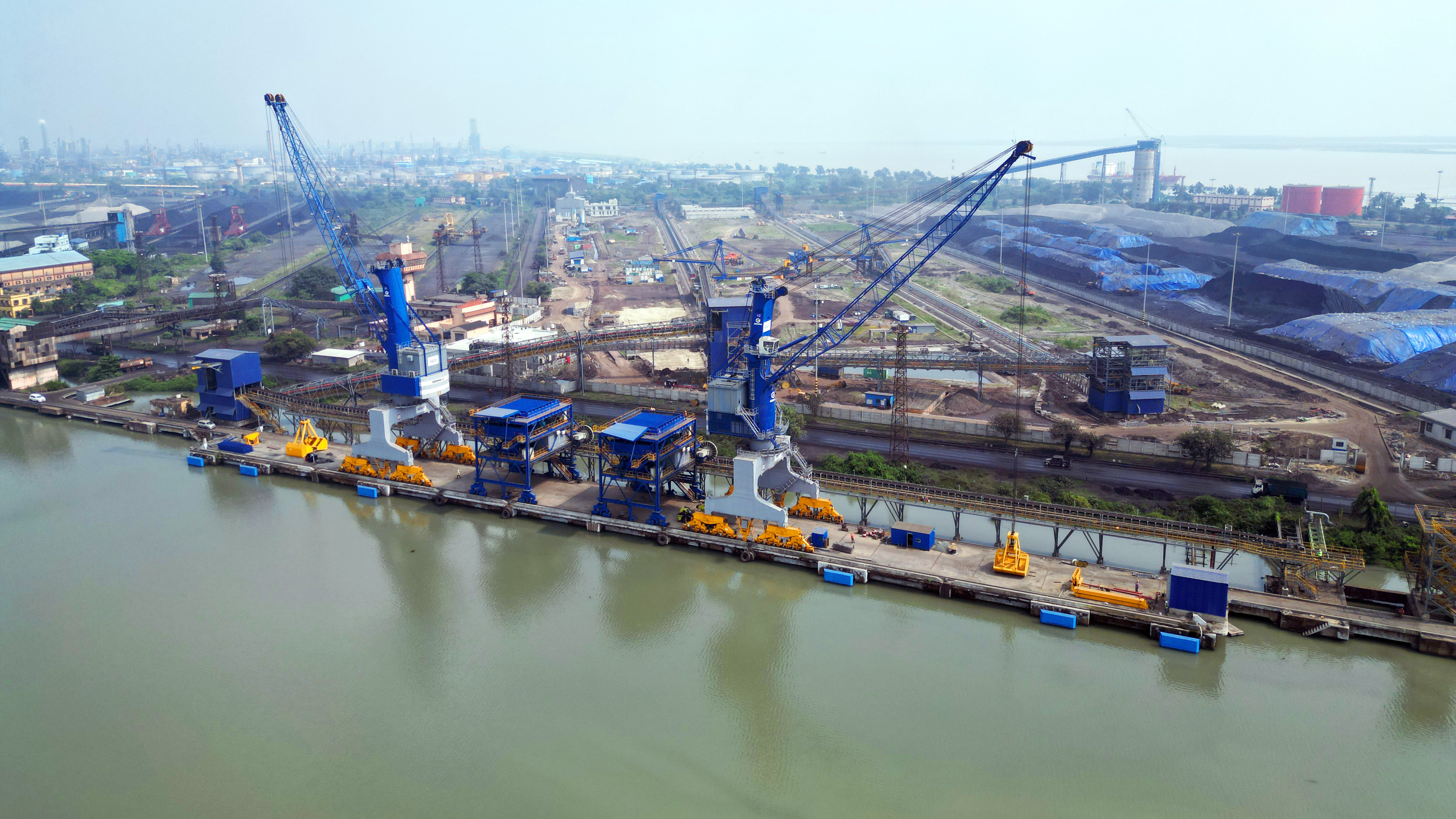 Aerial view of an industrial port with large blue cranes on the dock next to a river, coal piles in the background.
