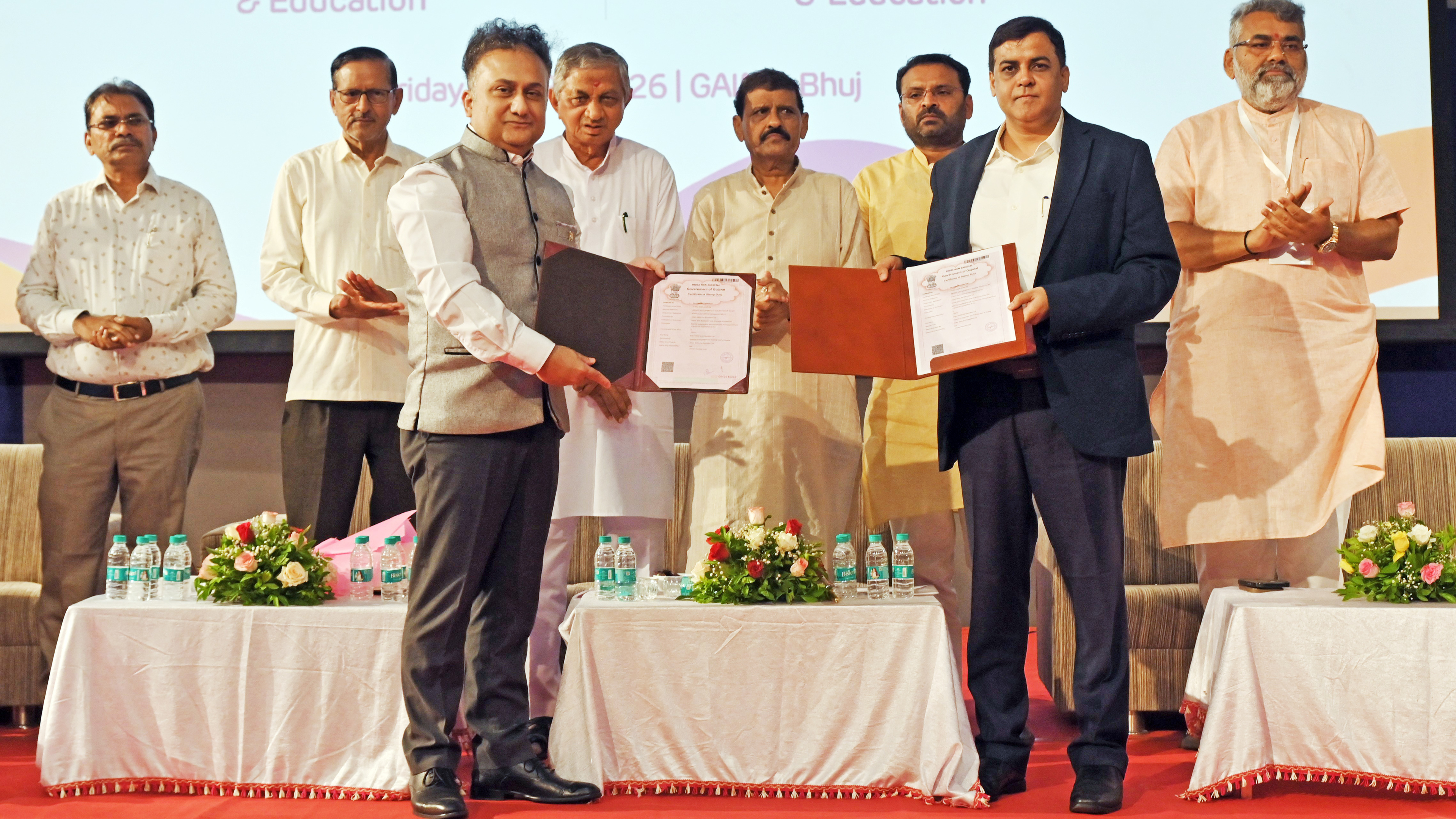 Two men in suits exchange signed documents on stage, while other men observe.